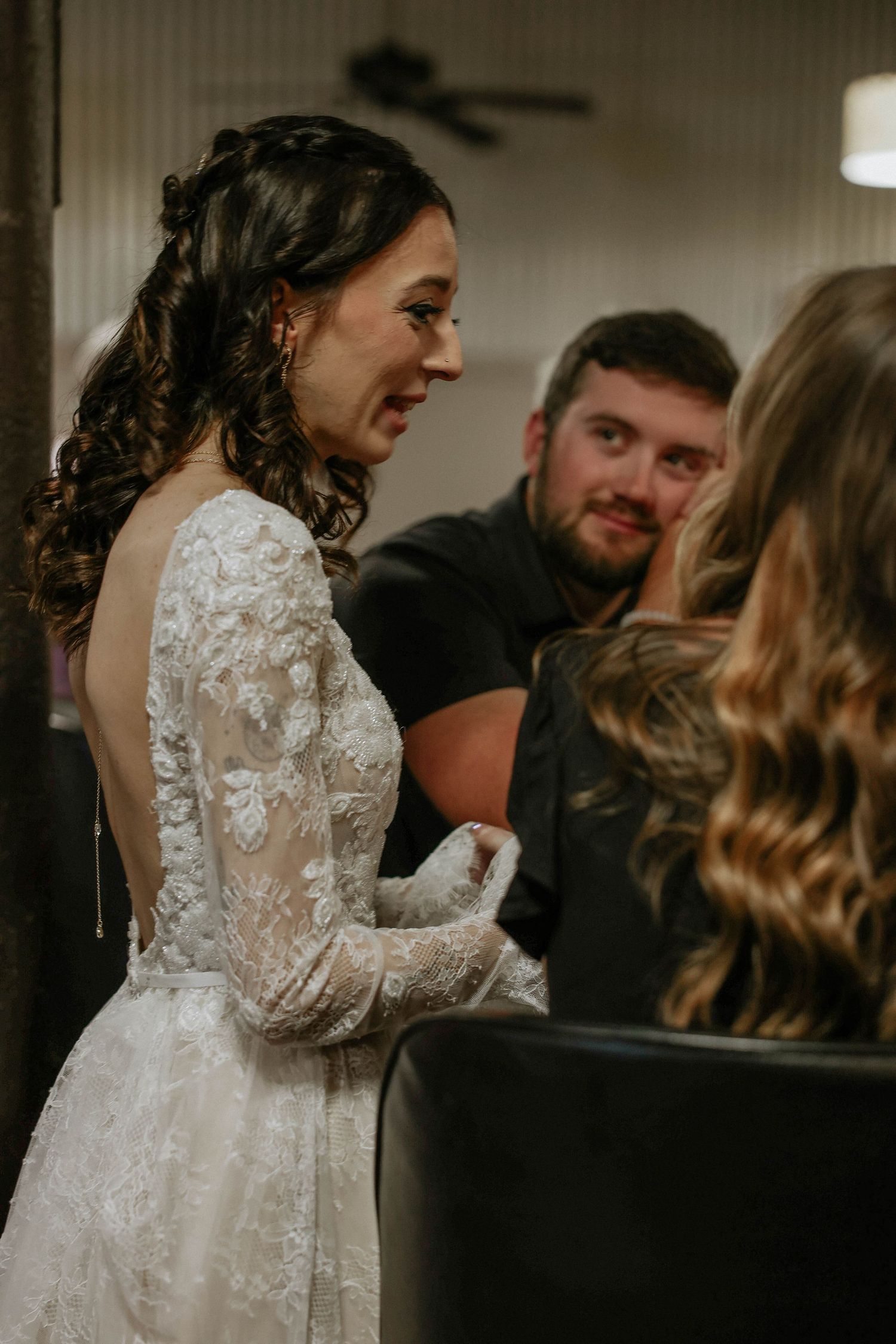 Bride in elegant lace long-sleeve backless wedding gown smiling and conversing with guests at wedding reception.