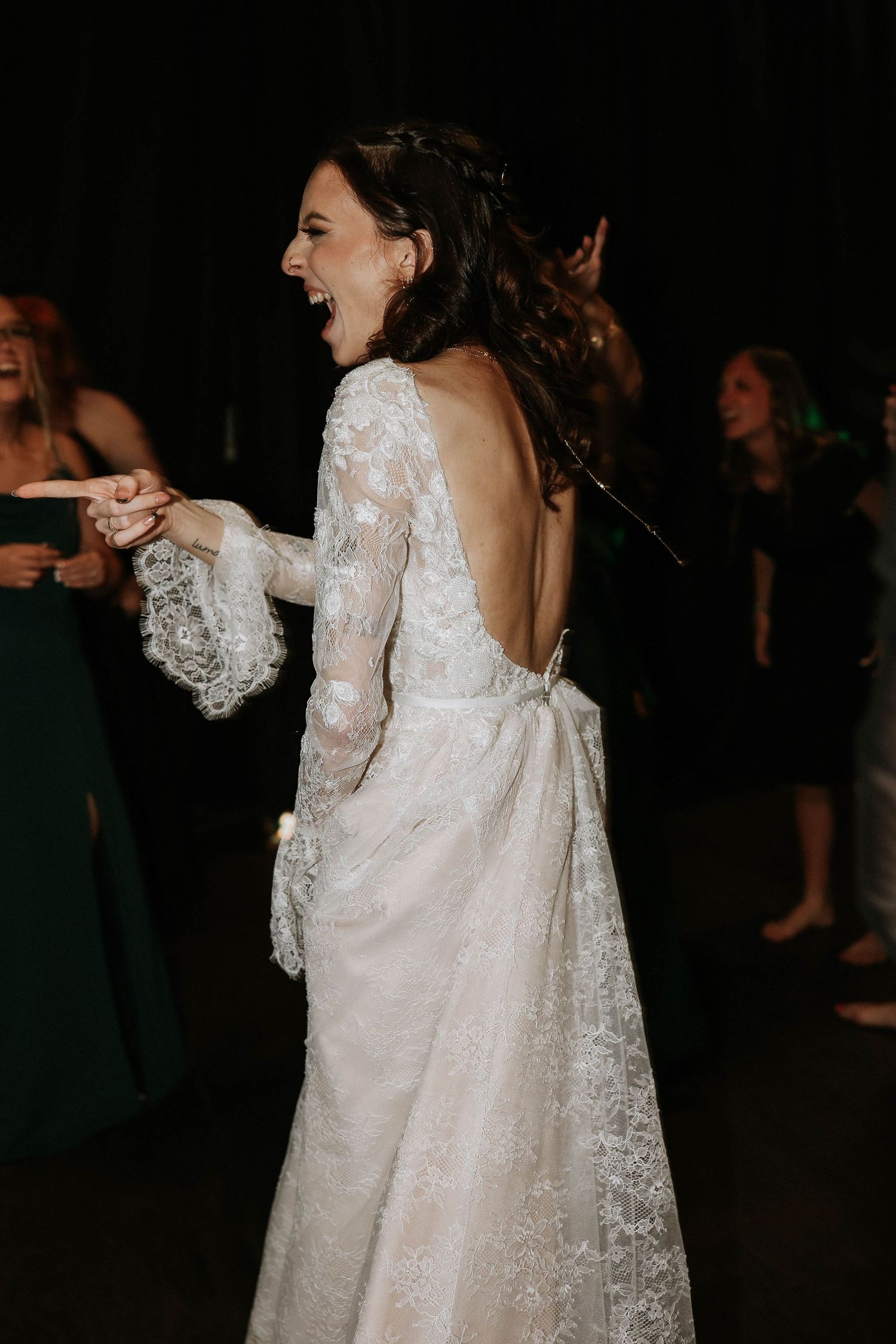 Bride in backless lace wedding gown with bow laughing joyfully while dancing at elegant wedding reception.