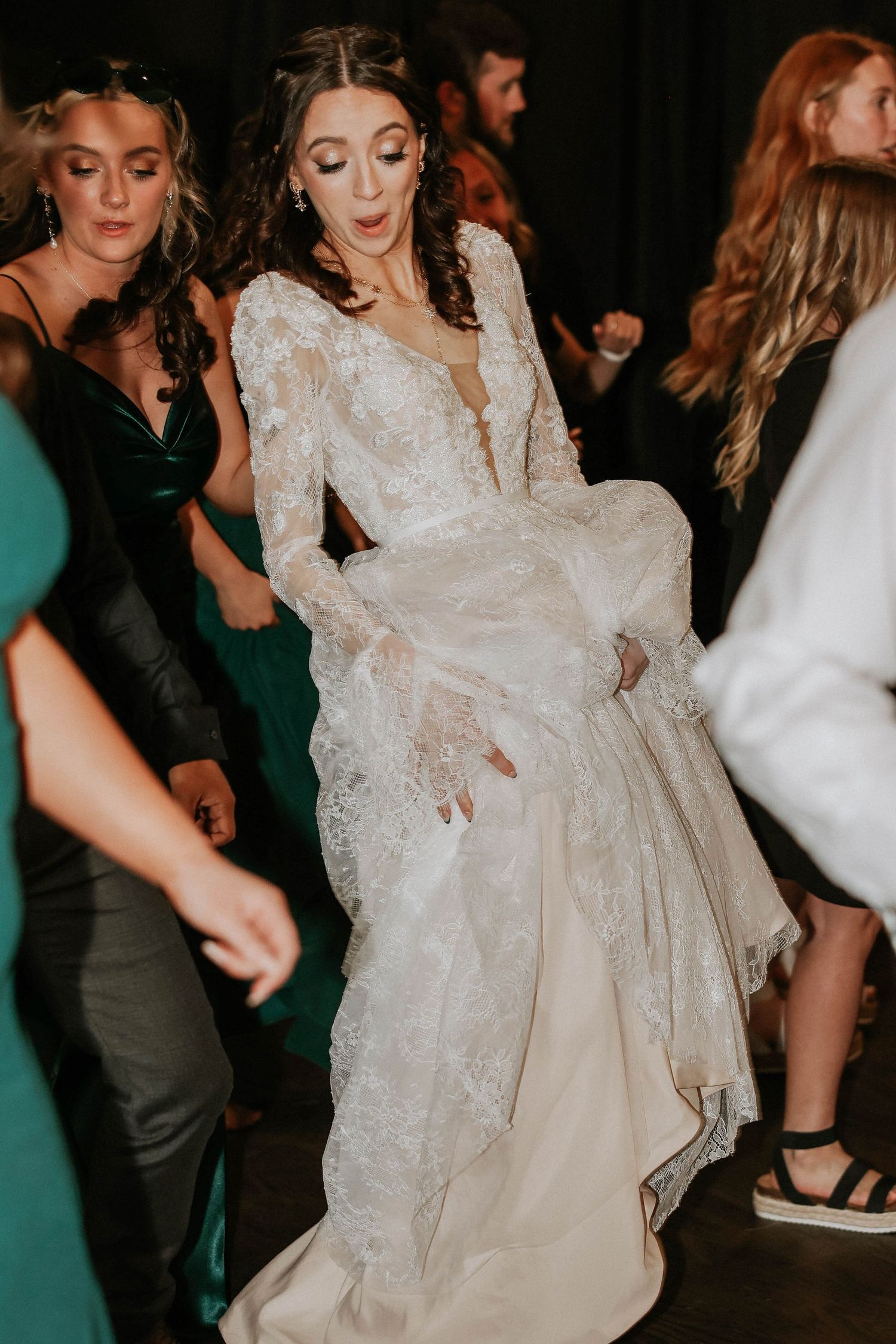 Bride in lace long-sleeve wedding gown dancing joyfully at reception surrounded by guests in formal attire.