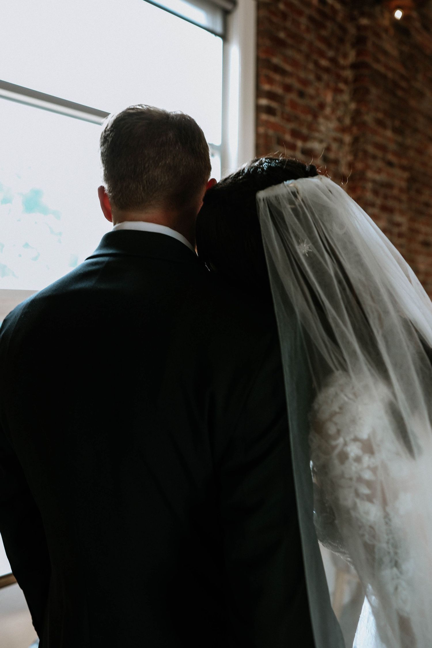 Bride and groom seen from behind, sharing an intimate moment near a window in a brick-walled venue.
