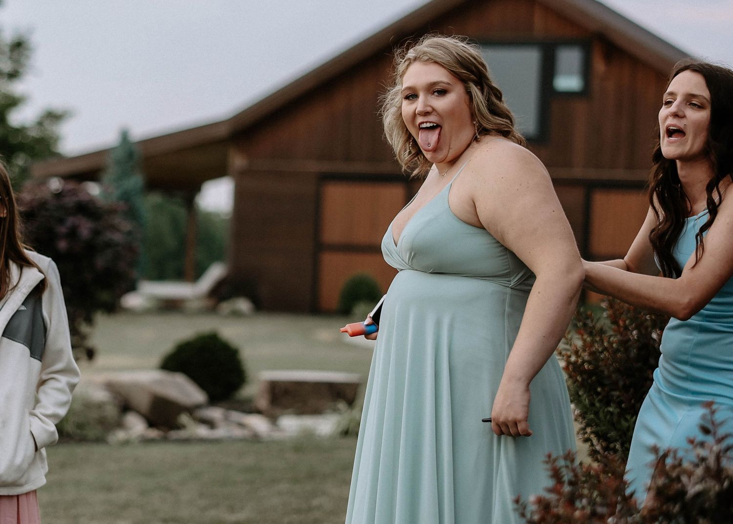 Two bridesmaids in light blue gowns laughing and celebrating outdoors near a rustic wooden venue.