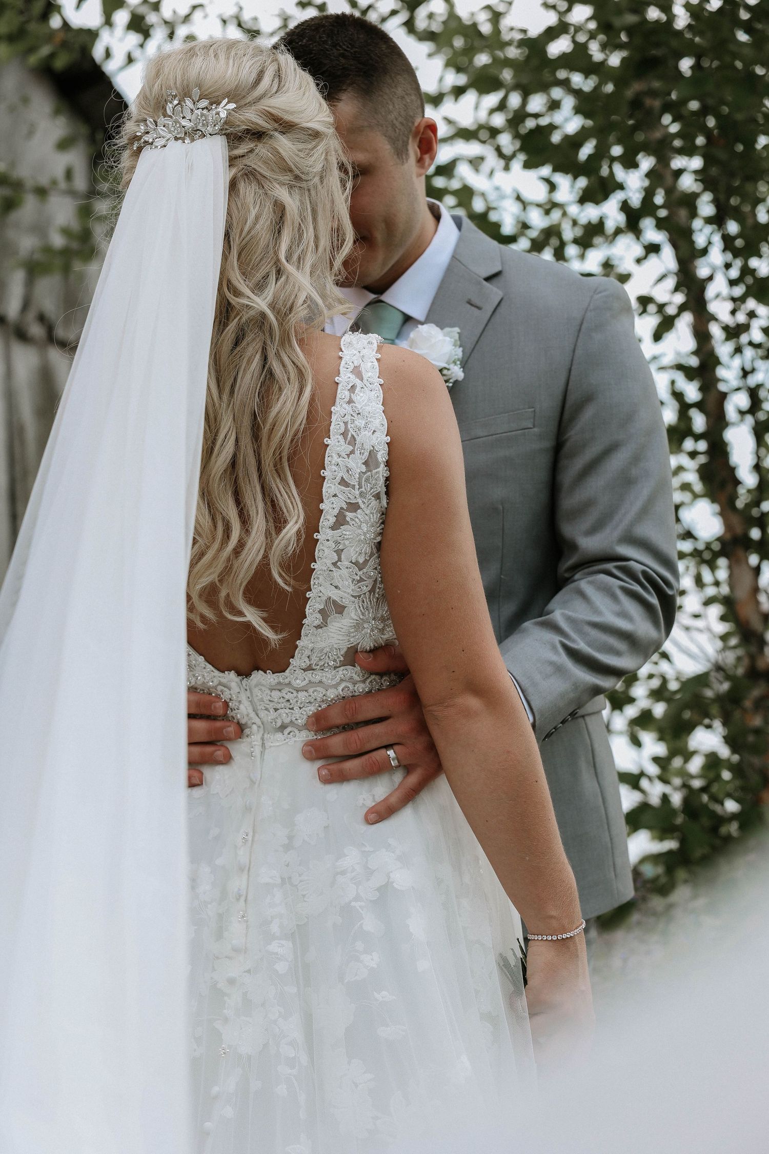 Groom in gray suit embraces bride in lace wedding dress with veil and hair accessory among green trees.