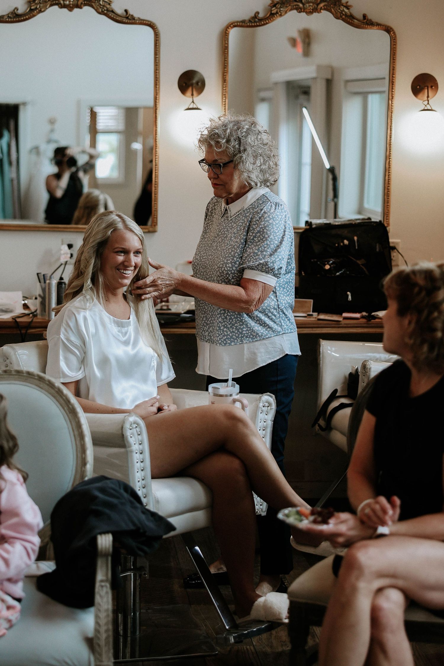 Bride smiling in a salon chair while a stylist applies makeup during wedding day preparations.