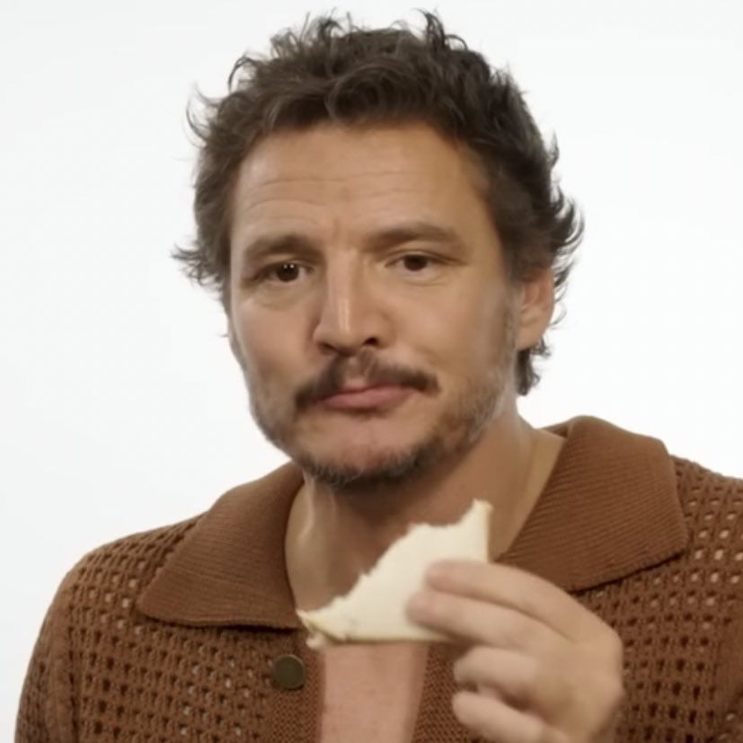 A man with a mustache and beard wearing a brown knit sweater holds a small piece of food against a white background.