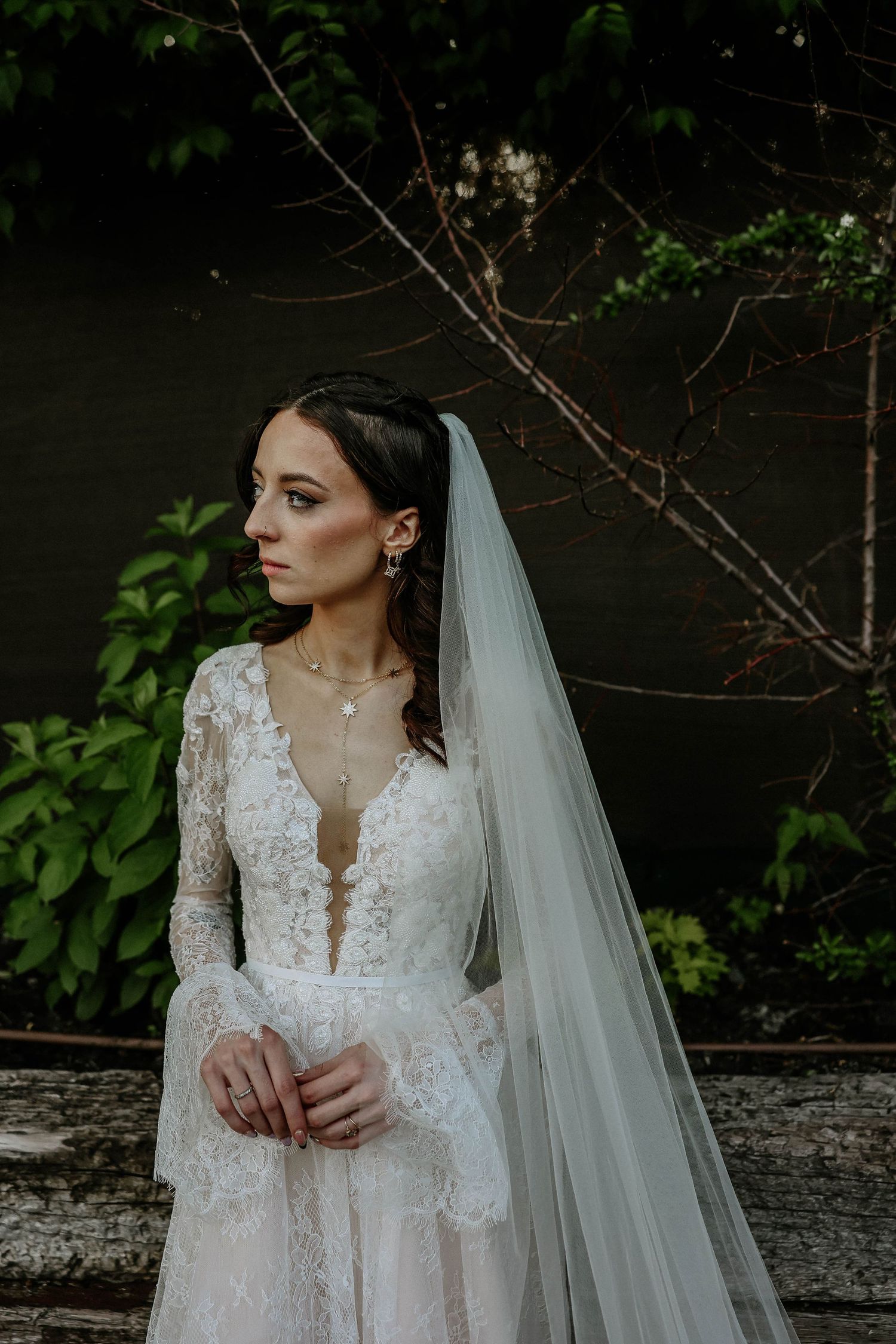 Bride in lace long-sleeve wedding gown with cathedral veil poses outdoors among lush green foliage.