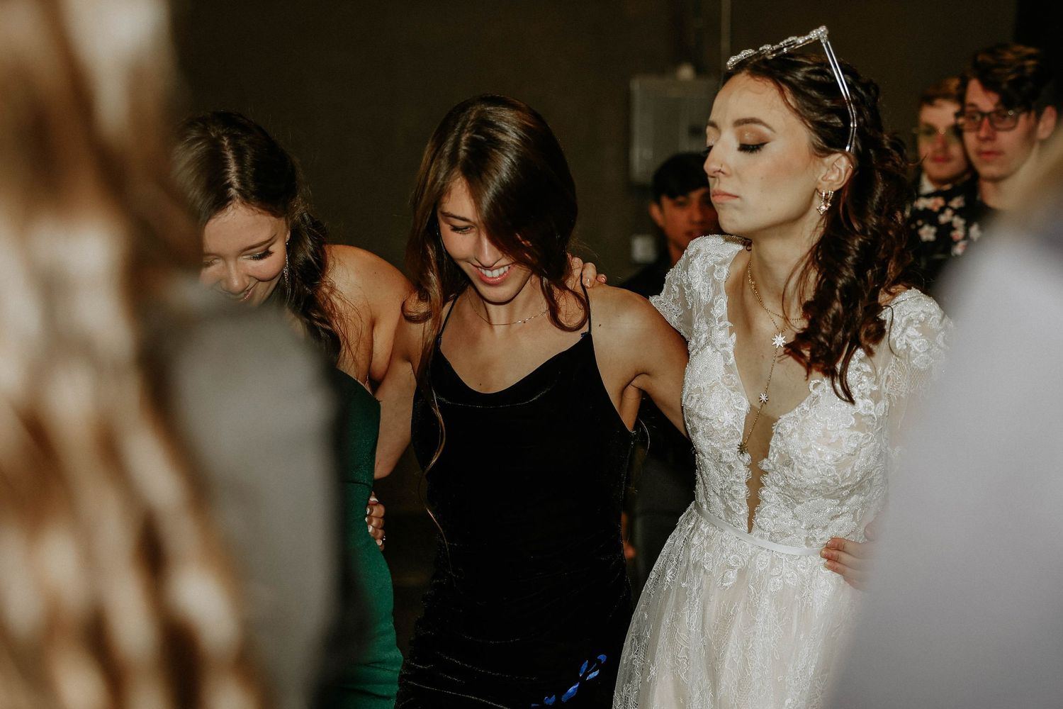 Bride in white lace gown dancing with bridesmaids in black and green dresses at a wedding reception.