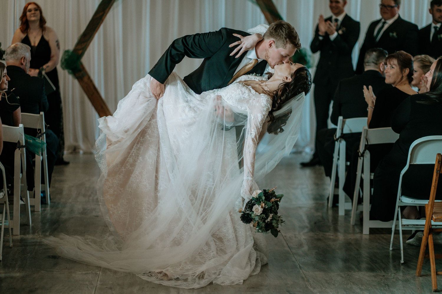 Groom dips bride in elegant white gown during romantic first dance at indoor wedding ceremony.