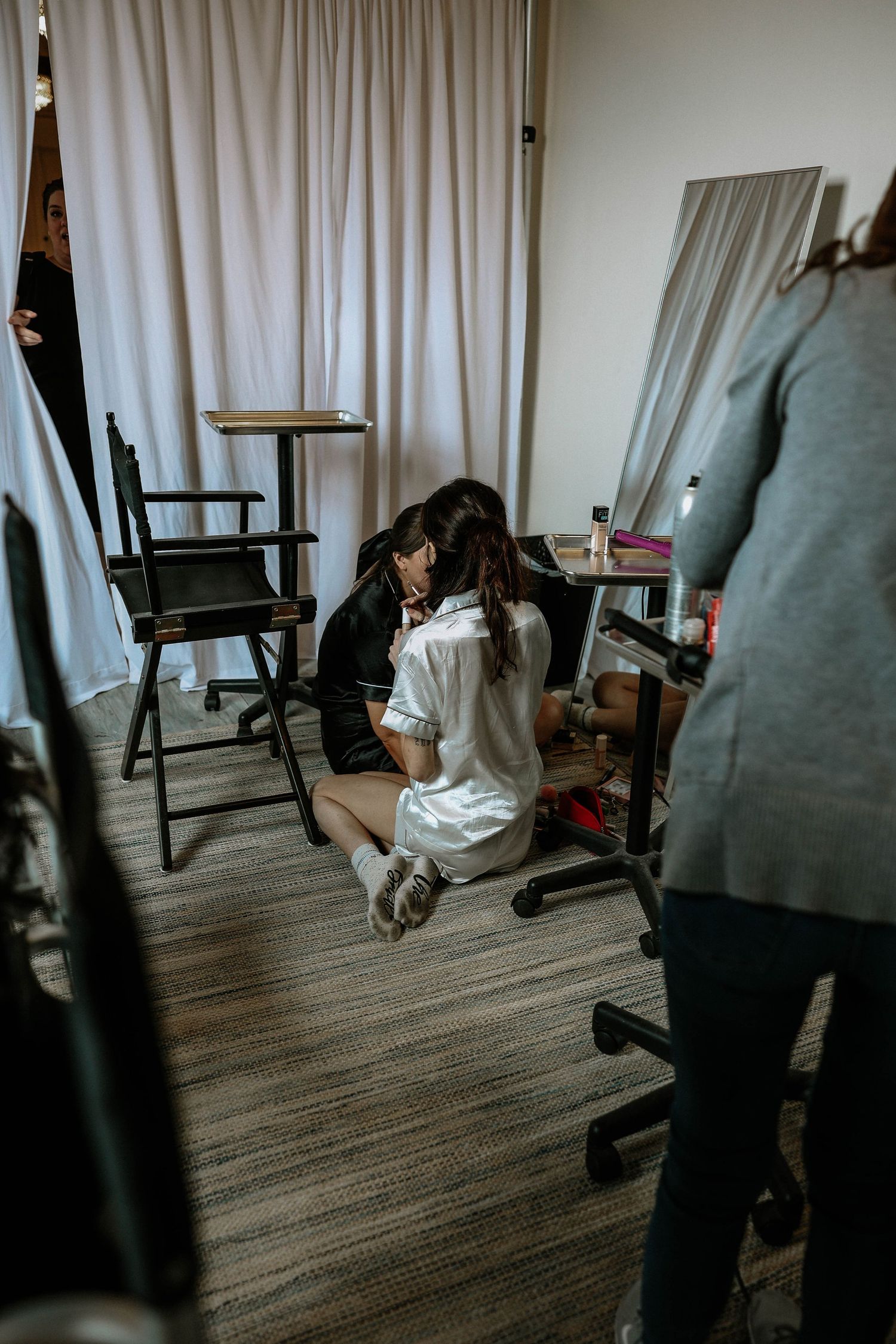 Two women in satin robes sit on the floor of a bridal suite during wedding day preparations surrounded by styling equipment.