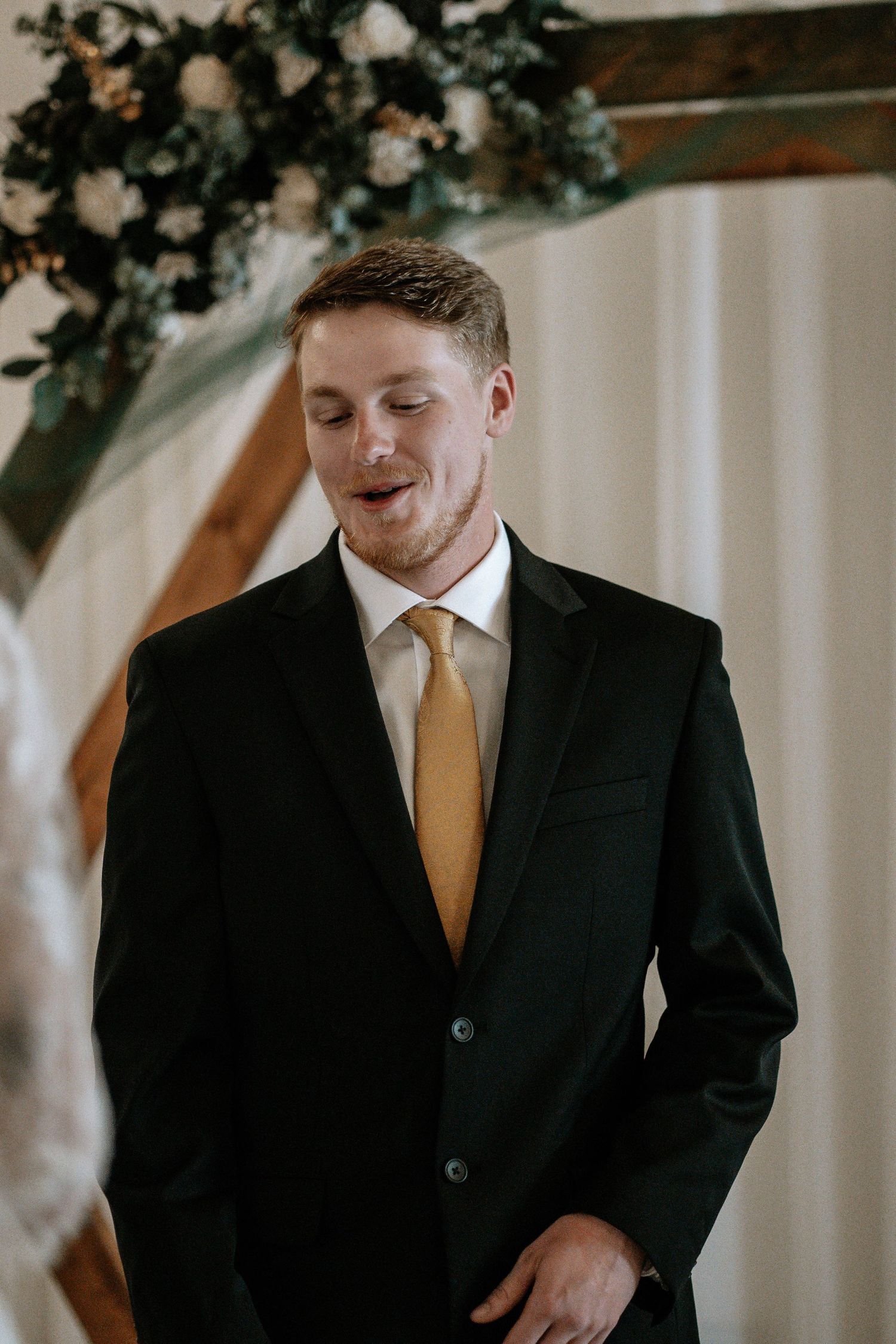 Groom in black suit and gold tie smiling emotionally at wedding ceremony altar with floral arch.