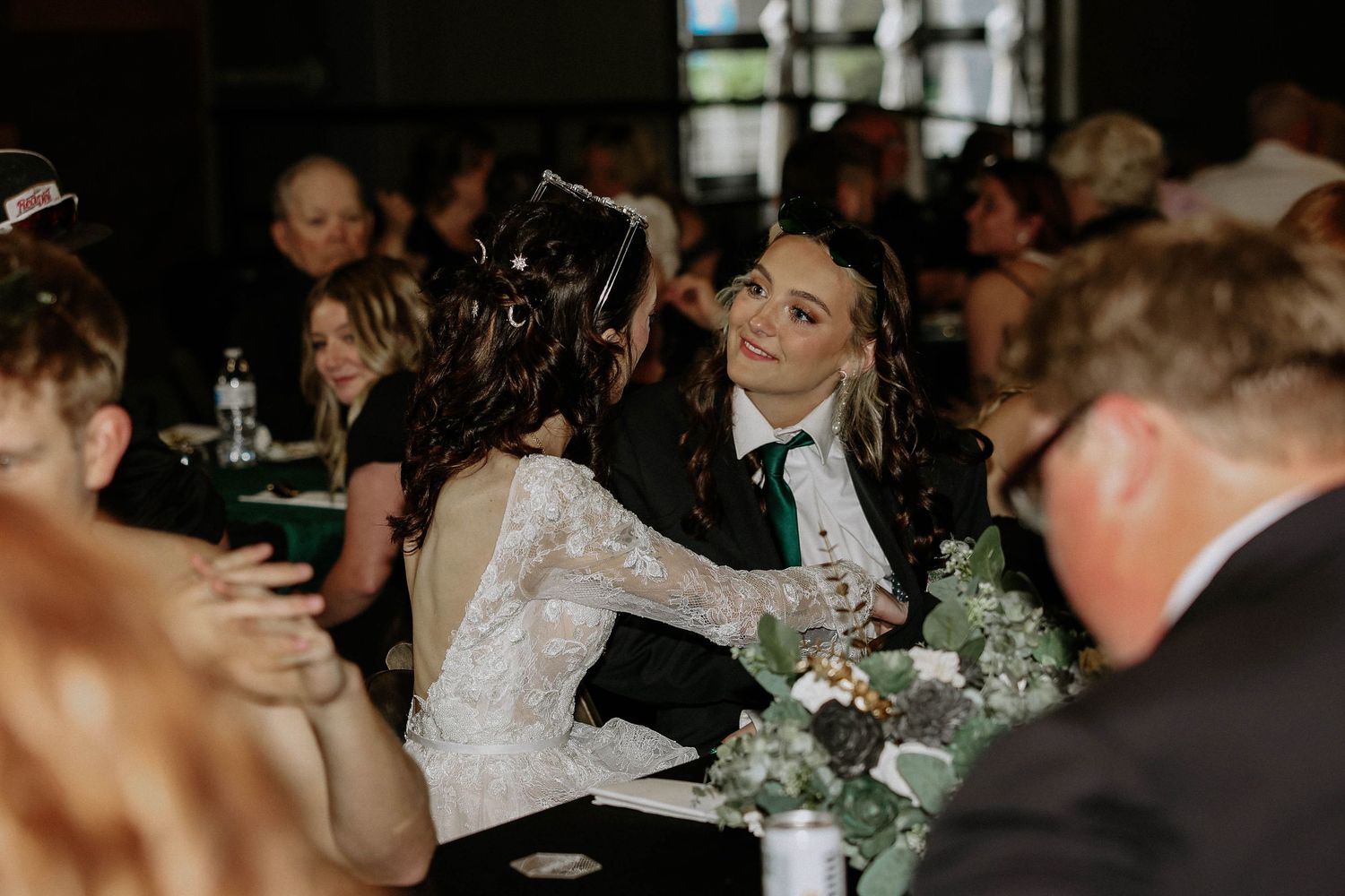 Two brides share a joyful moment at their wedding reception, one in white lace, the other in a black tuxedo with green tie.