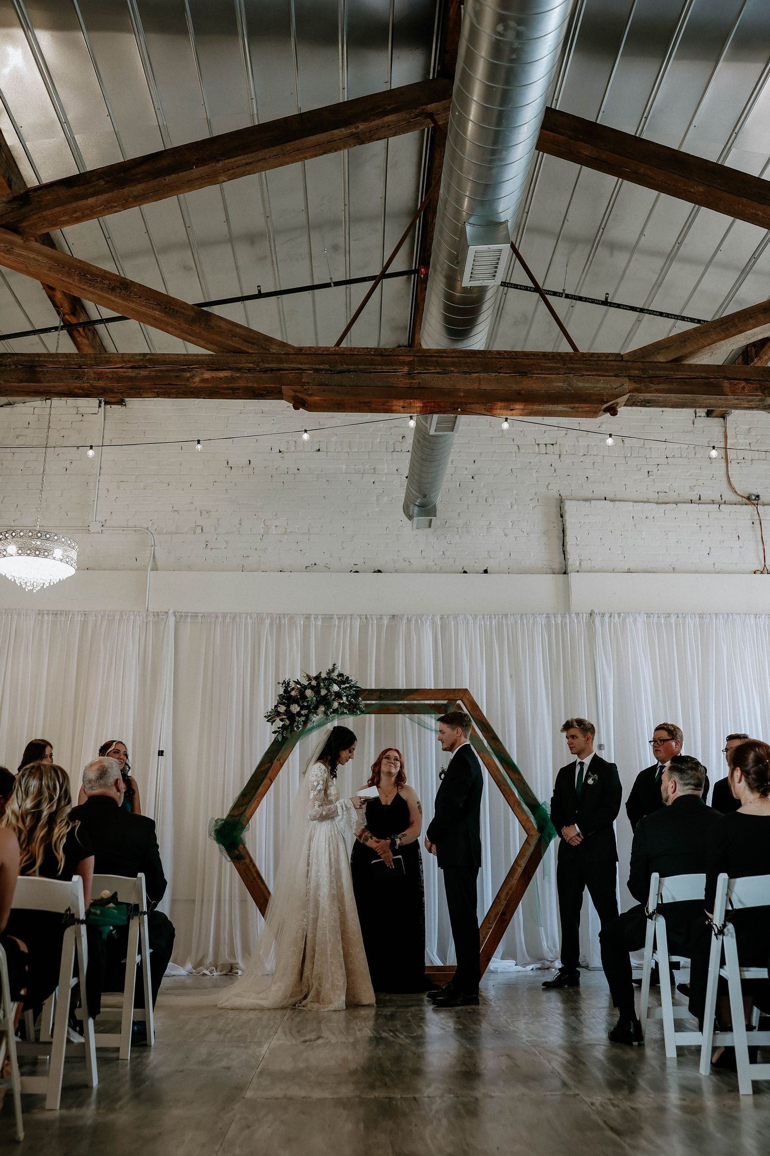 Wedding ceremony in industrial venue with hexagonal wooden arch, white draping, exposed beams, bride and groom exchanging vows.