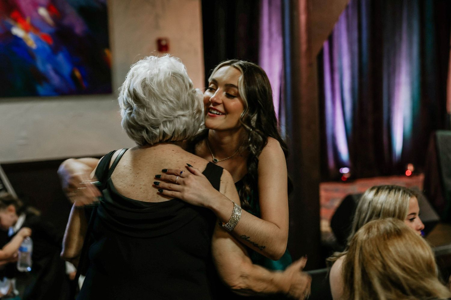 Two women sharing a warm hug at a formal event, smiling joyfully in elegant black attire.