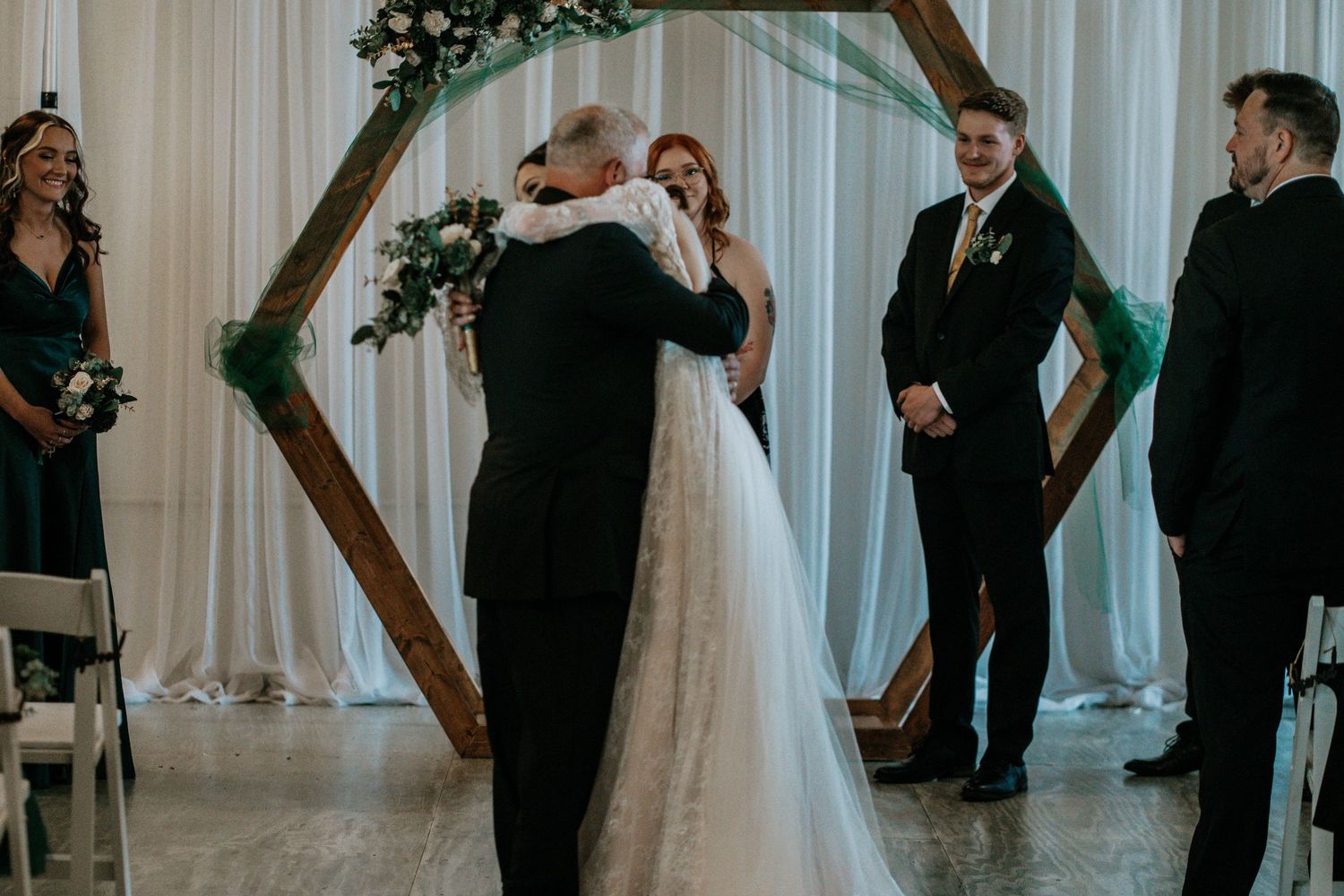 Bride hugging her father at the altar during an indoor wedding ceremony with a hexagonal wooden arch.