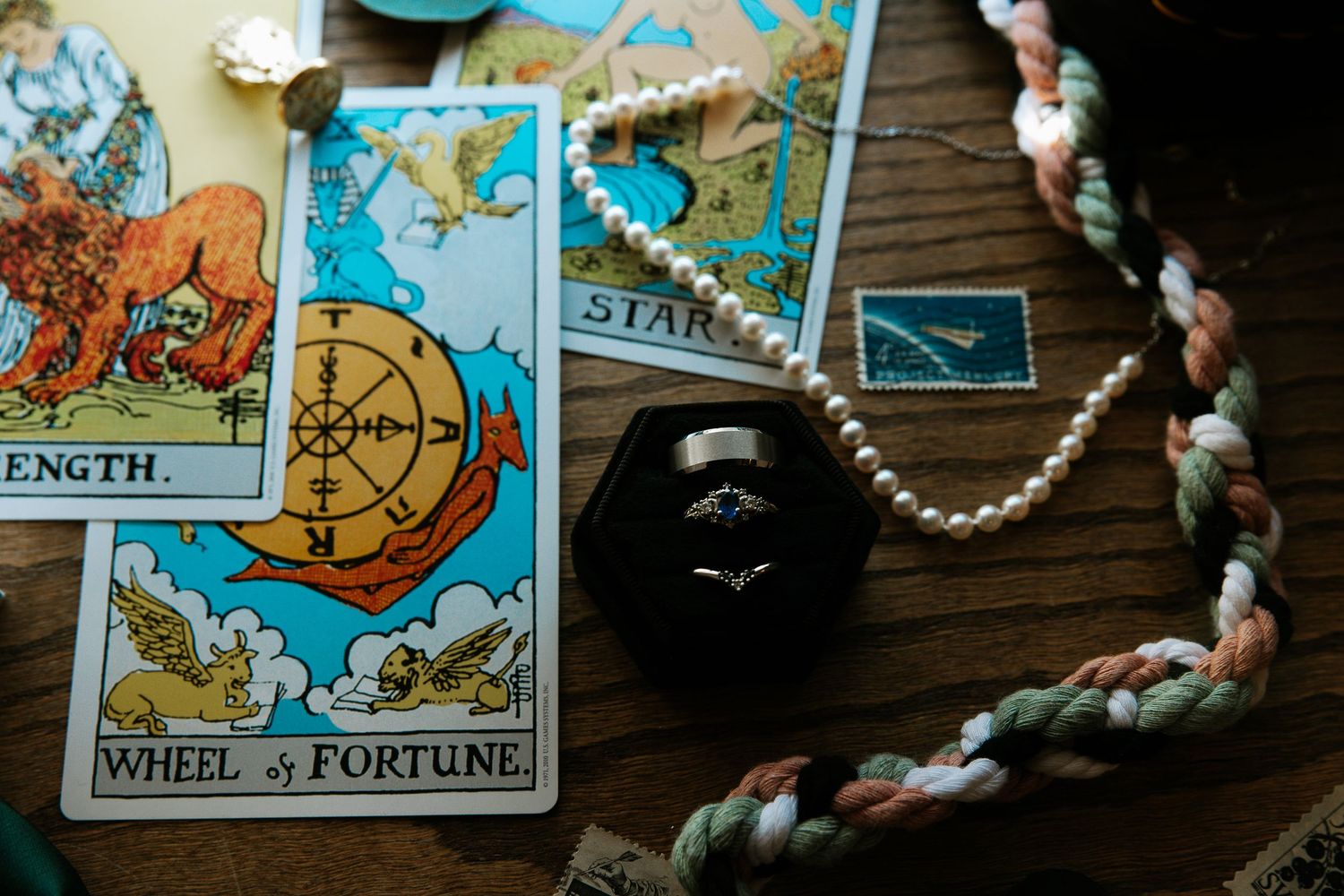 Tarot cards with diamond ring in black box, pearl necklace, and braided cord on wood table.