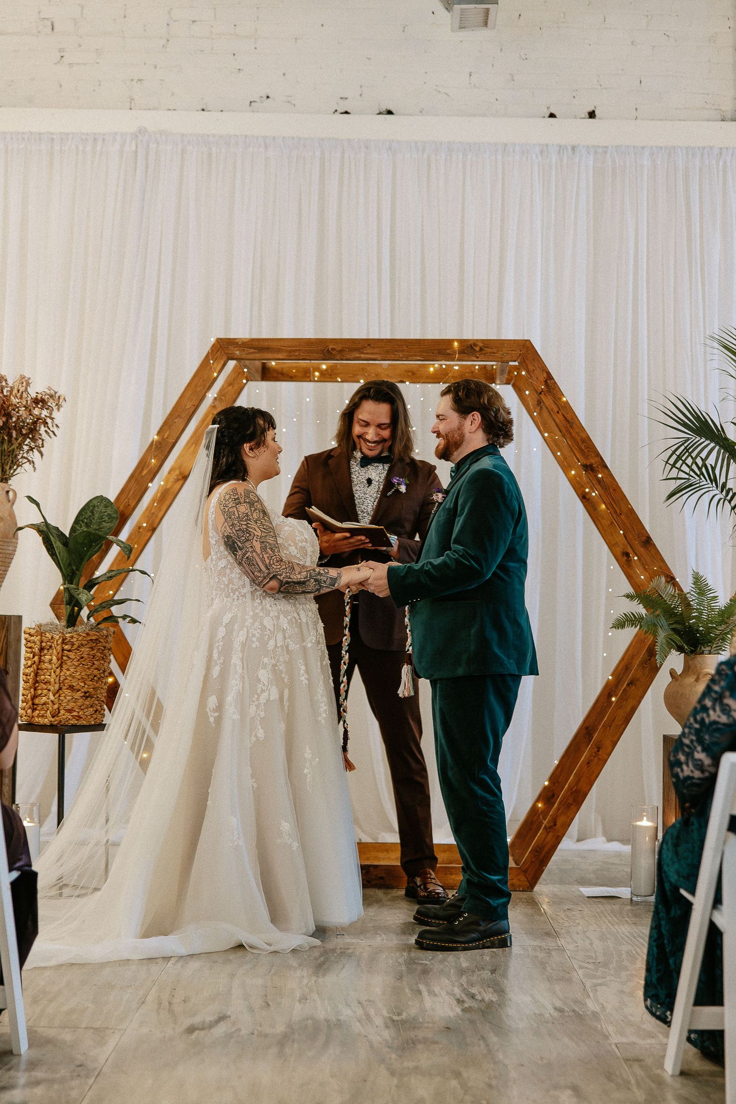 Bride and groom laugh with officiant during wedding ceremony at hexagonal wooden arch.