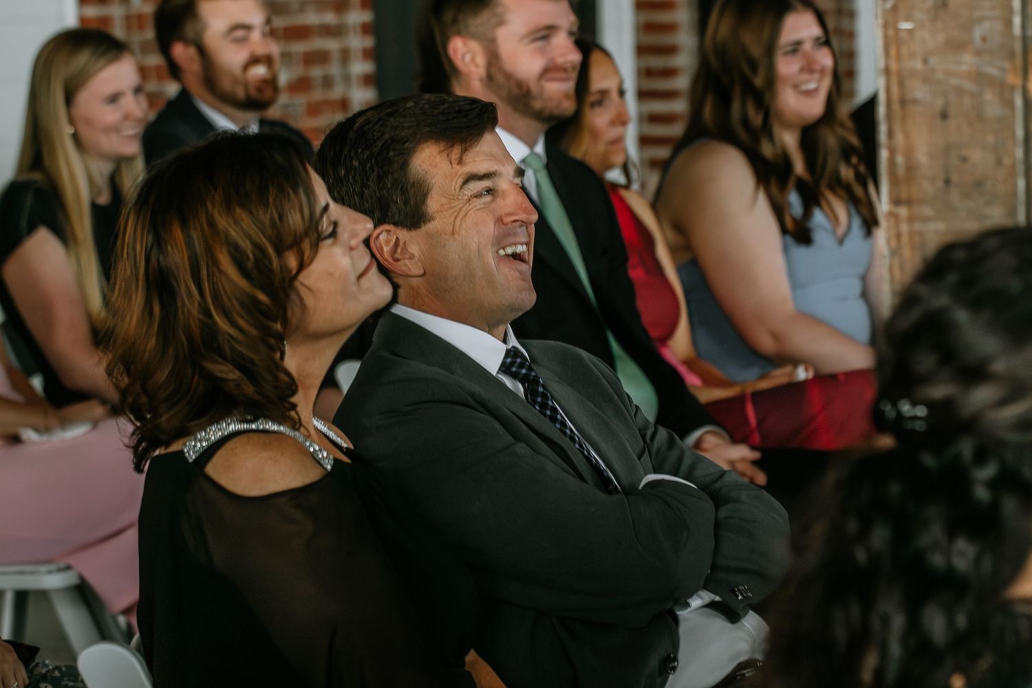 Couple laughing joyfully together seated among wedding guests during ceremony.