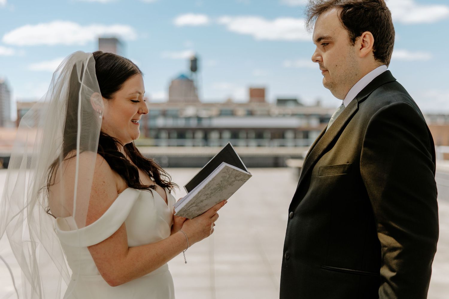 Bride reads wedding vows from a book while groom listens on a rooftop with city skyline backdrop.