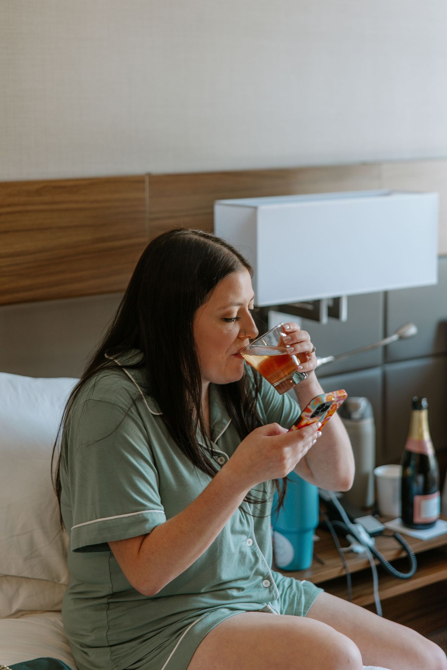 Woman in green pajamas sitting on bed drinking from a glass beside a champagne bottle.