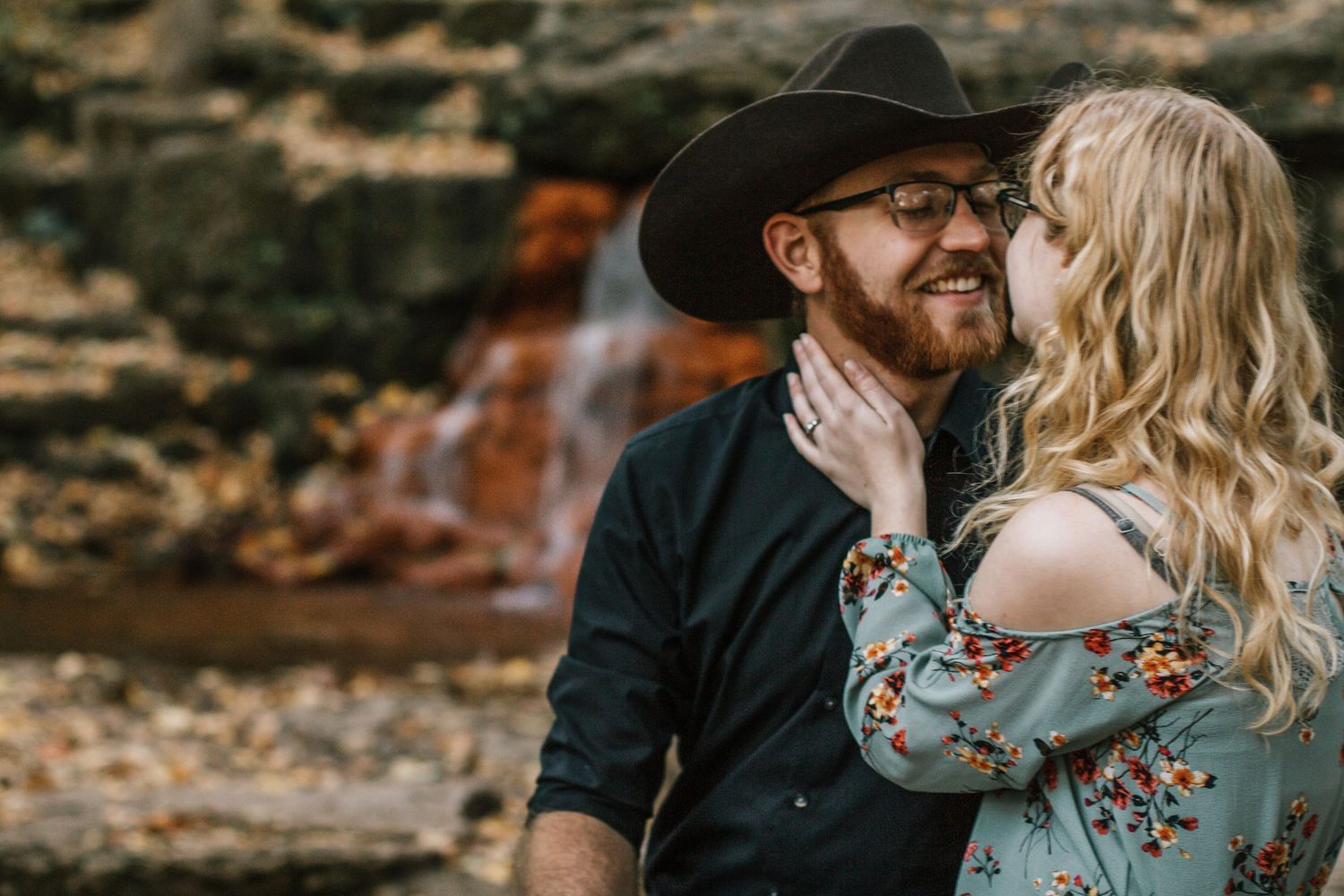 A smiling bearded man in a black hat and glasses receives a gentle kiss from a woman near a waterfall in Yellow Springs, Ohio