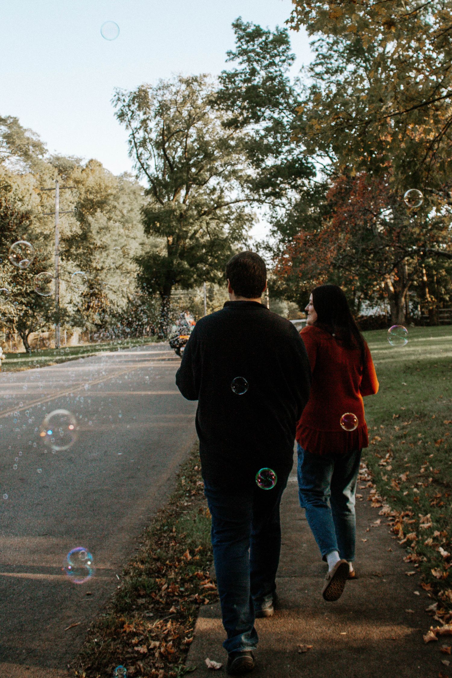 Couple walking on a park path surrounded by floating bubbles during an autumn engagement photo session in Yellow Springs, Ohio