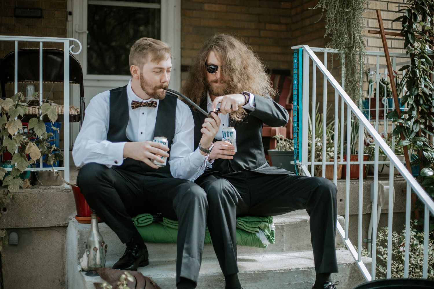 Two groomsmen in black tuxedos and bow ties sit on front steps sharing drinks before a wedding in Cincinnati, Ohio