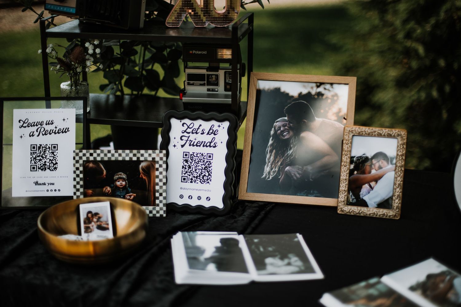 Outdoor wedding display table with framed photos, QR code signs, and Polaroids on a black tablecloth.