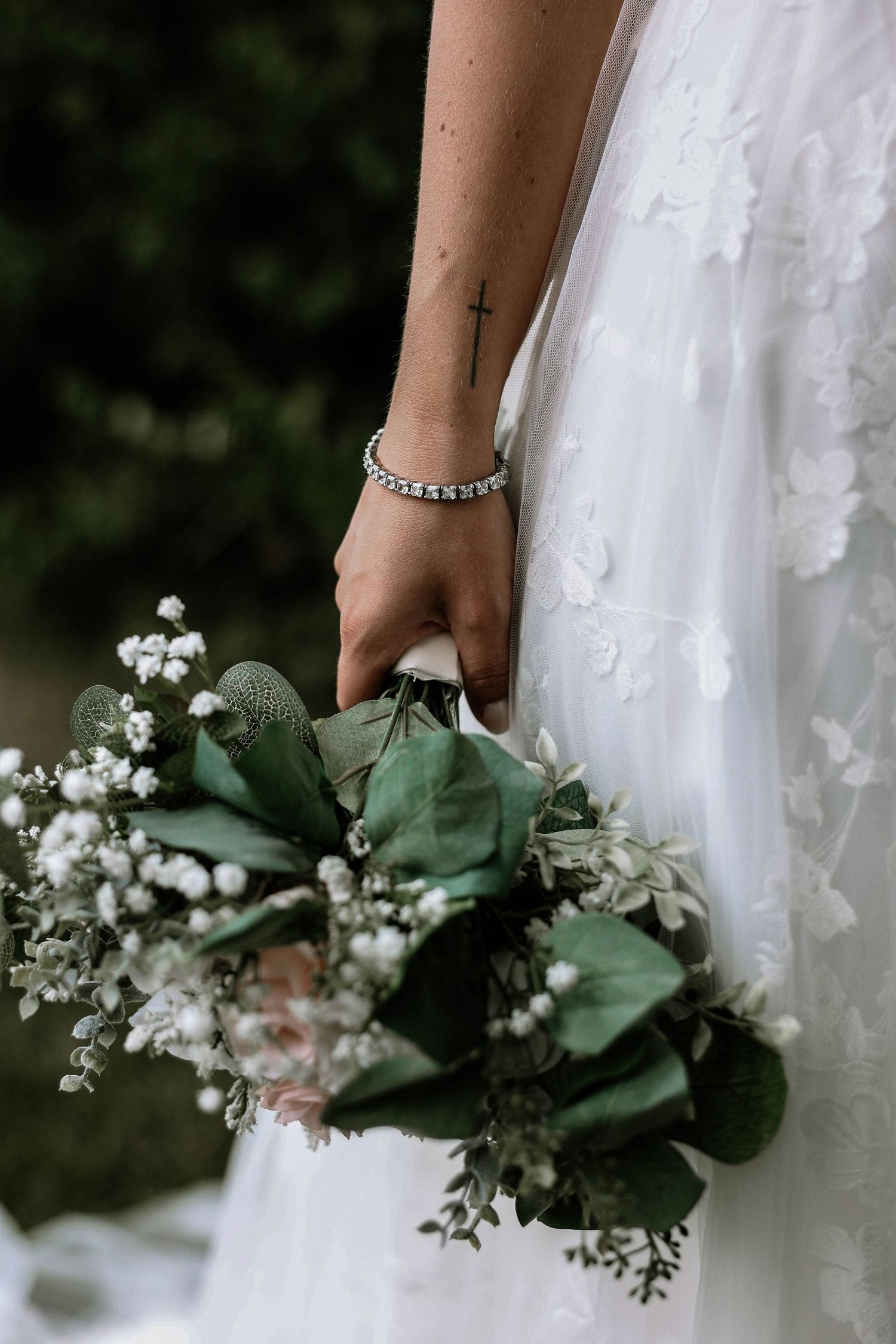 Bride in lace wedding gown holding a green and white bouquet with a cross tattoo on her wrist.