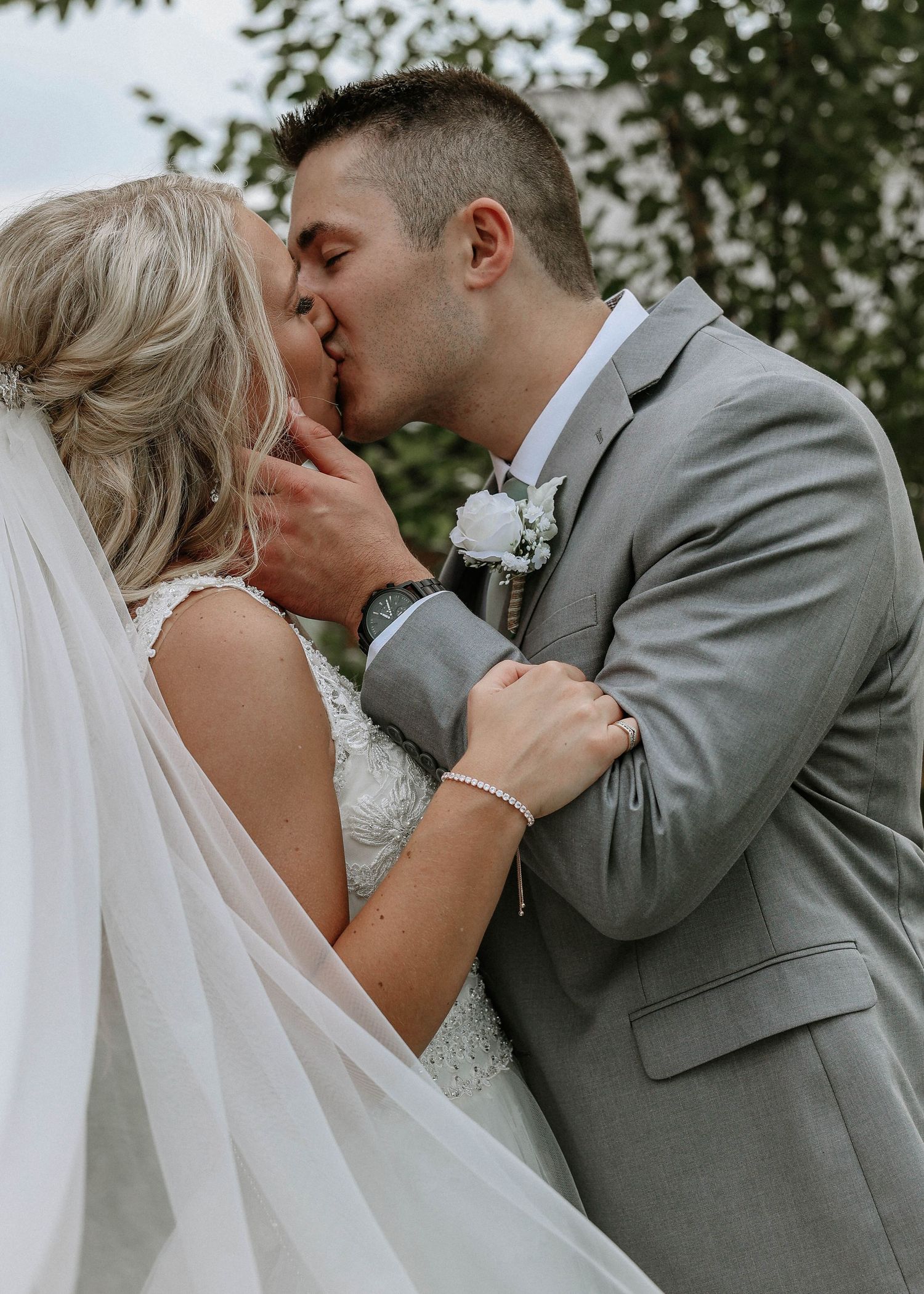 A bride and groom share a romantic kiss outdoors, with the bride in a lace gown and veil and groom in a gray suit.