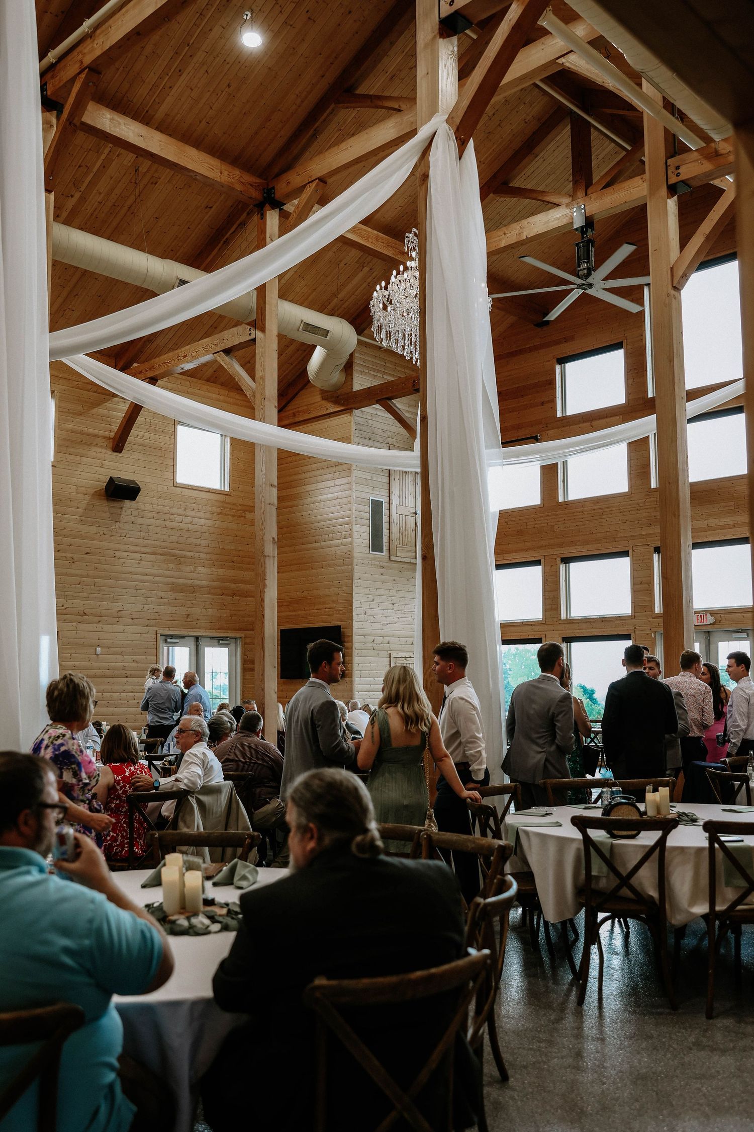 Rustic wedding reception venue with vaulted wood ceiling, draped fabric, chandelier, and seated guests.