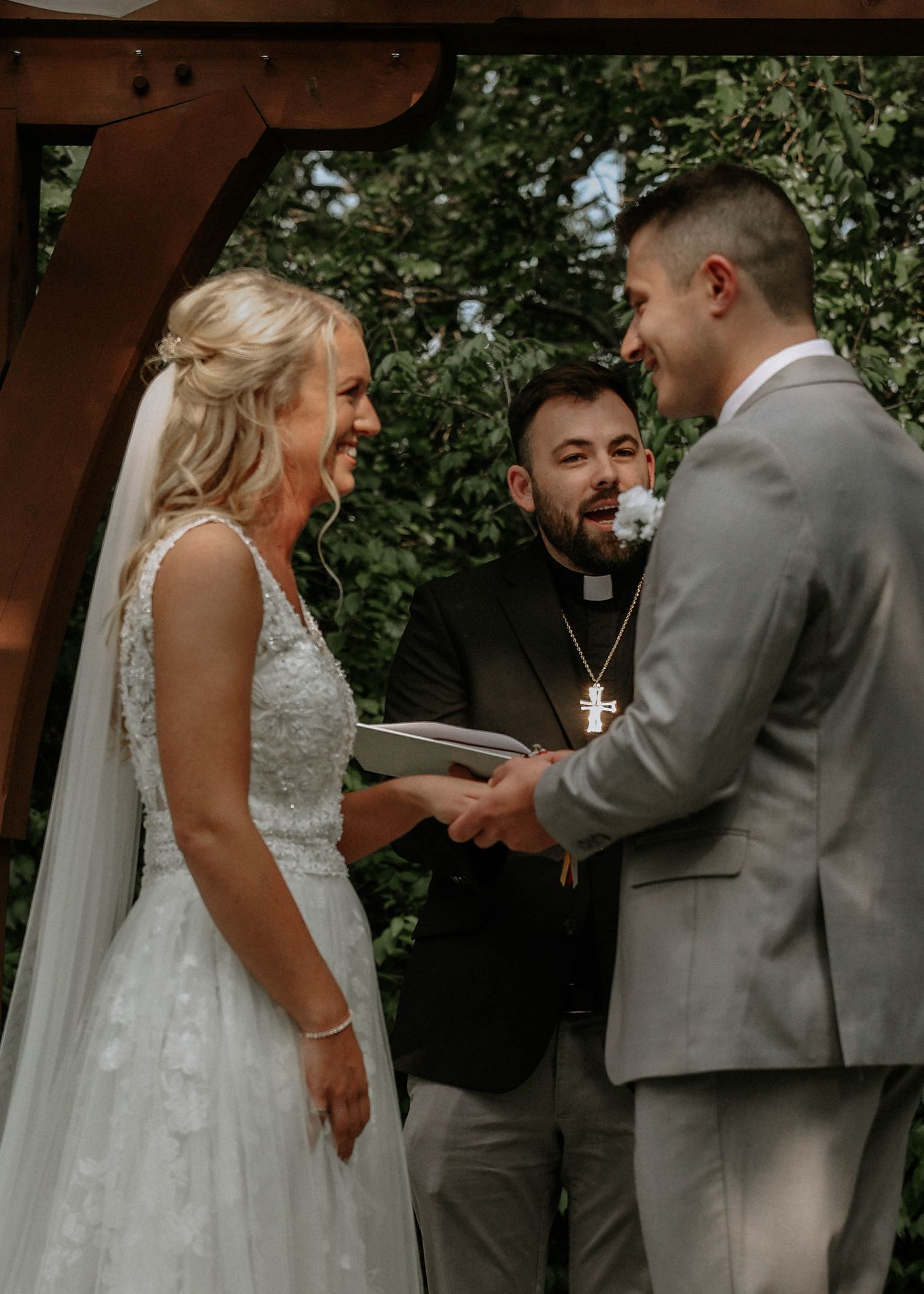 Bride and groom exchange vows outdoors under wooden arch with officiant wearing cross necklace at garden wedding ceremony.