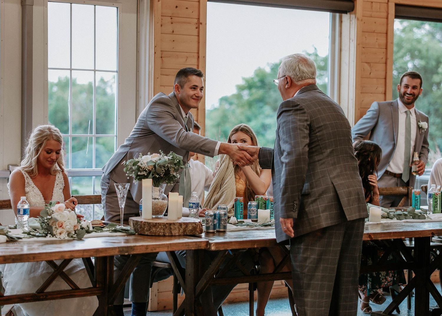 Groom in gray suit shaking hands with elderly guest at wedding head table during reception.