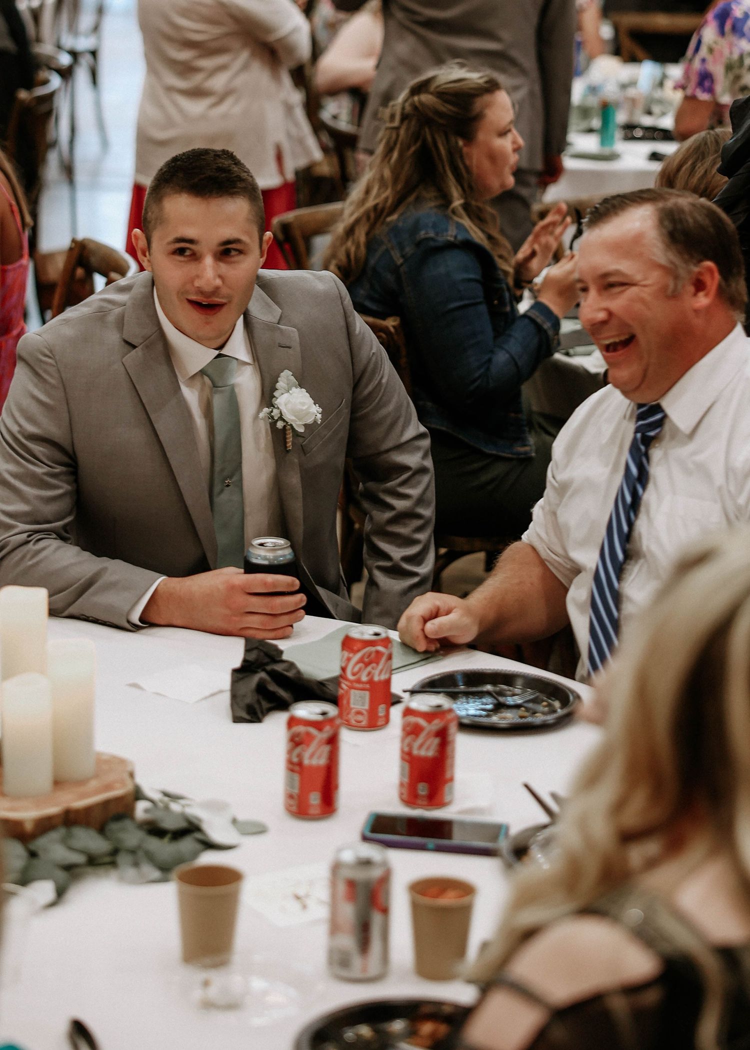 Young groom in gray suit laughing with older man at wedding reception table with Coca-Cola cans.