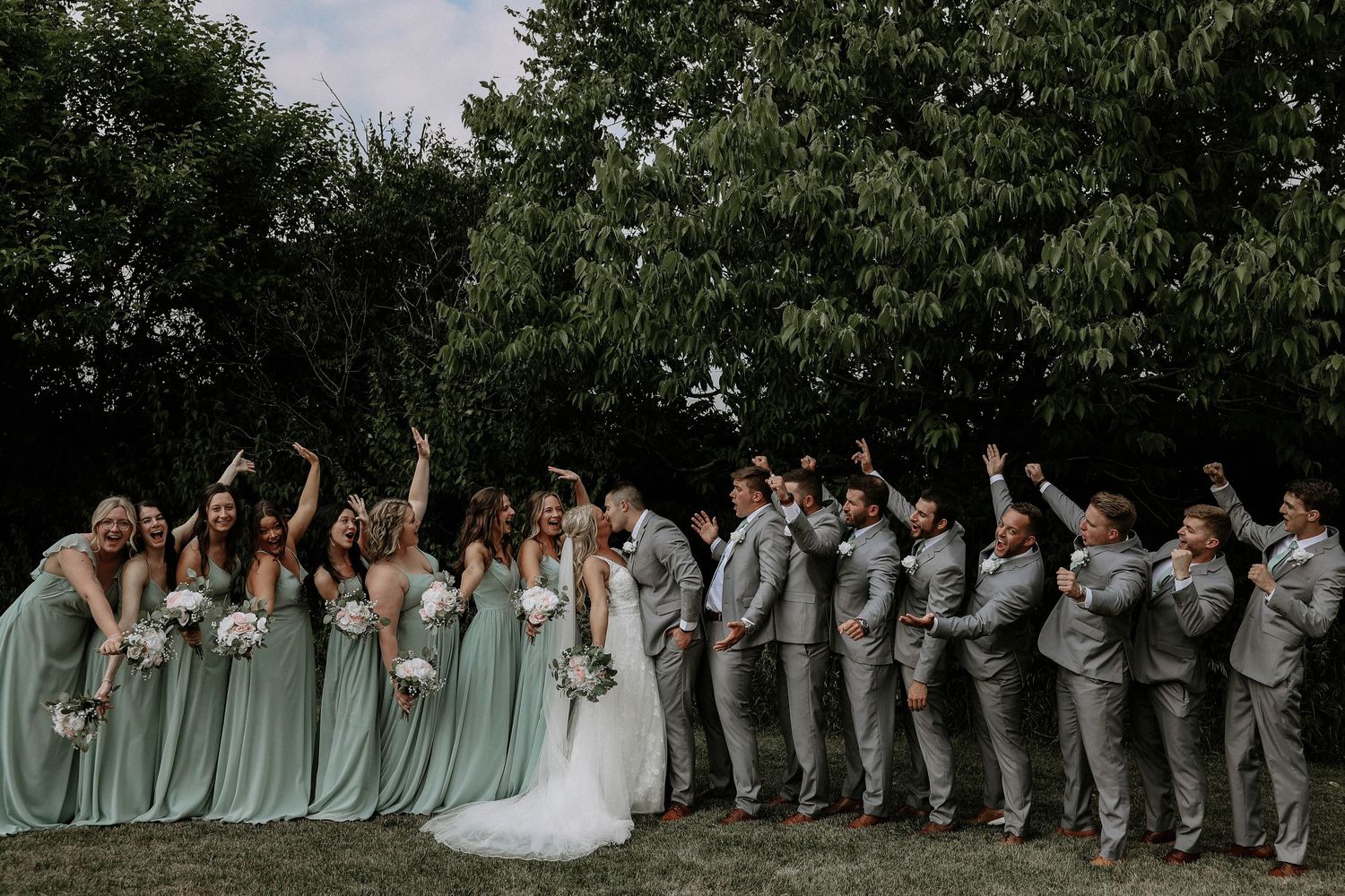 Wedding party celebrating outdoors, bridesmaids in sage green gowns and groomsmen in gray suits cheering with arms raised.