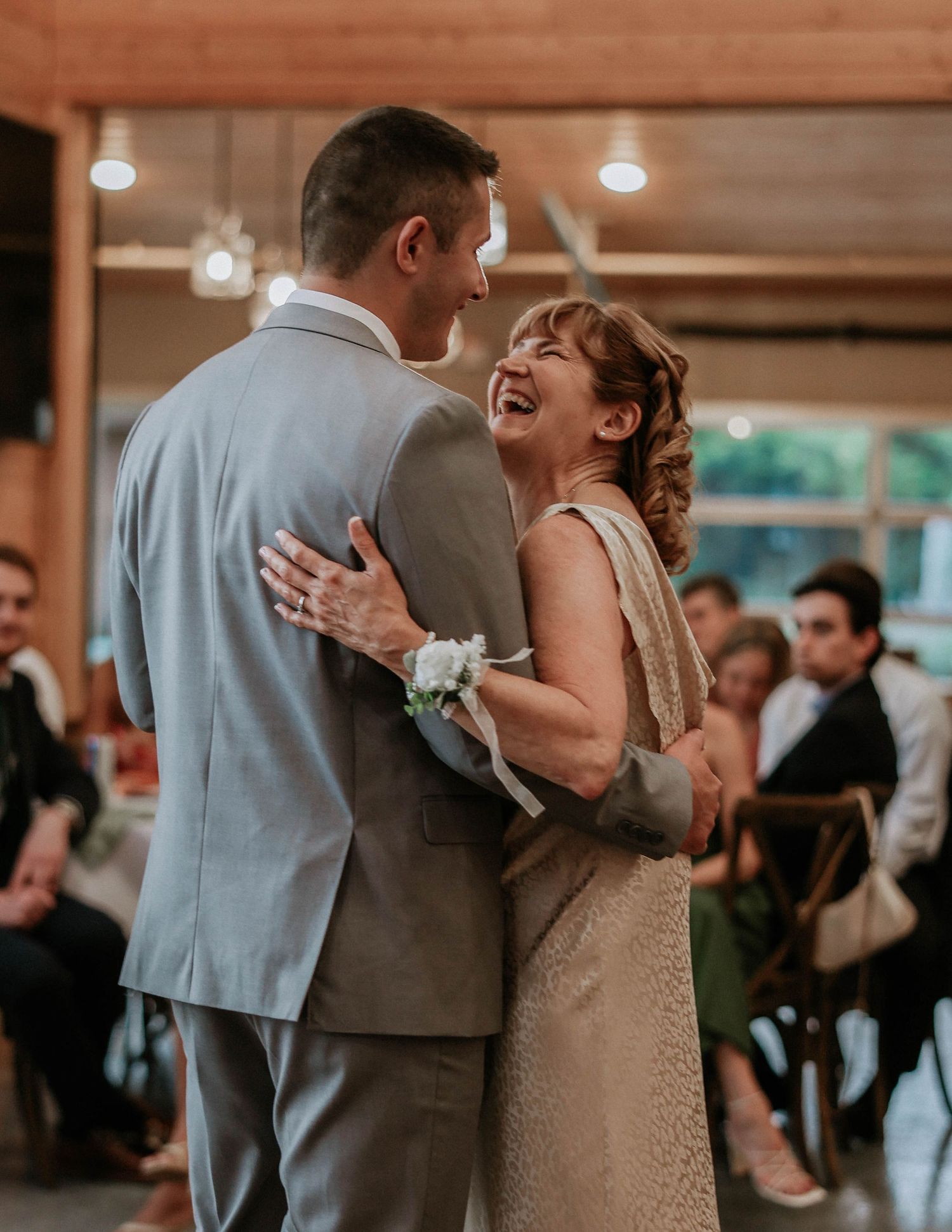 A joyful mother and son share a wedding dance, with the woman laughing warmly in a beaded gown with wrist corsage.