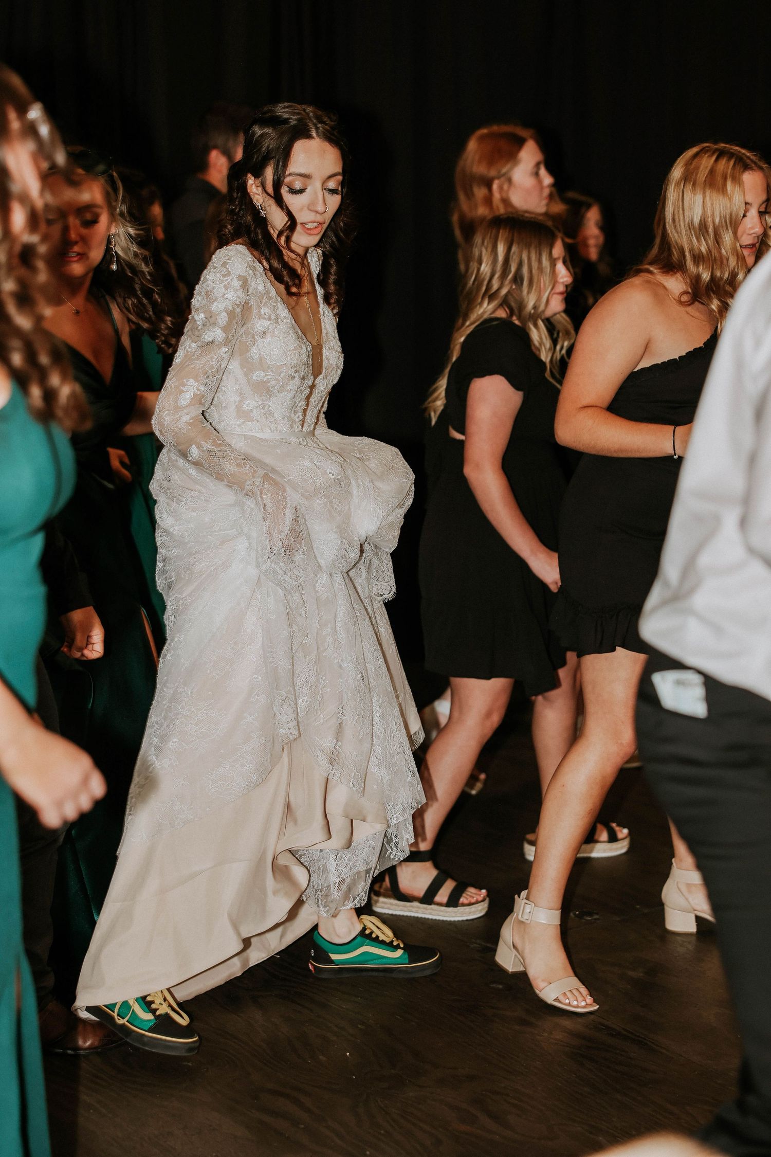 Bride in lace wedding gown and green Vans sneakers dancing joyfully with guests at a wedding reception.