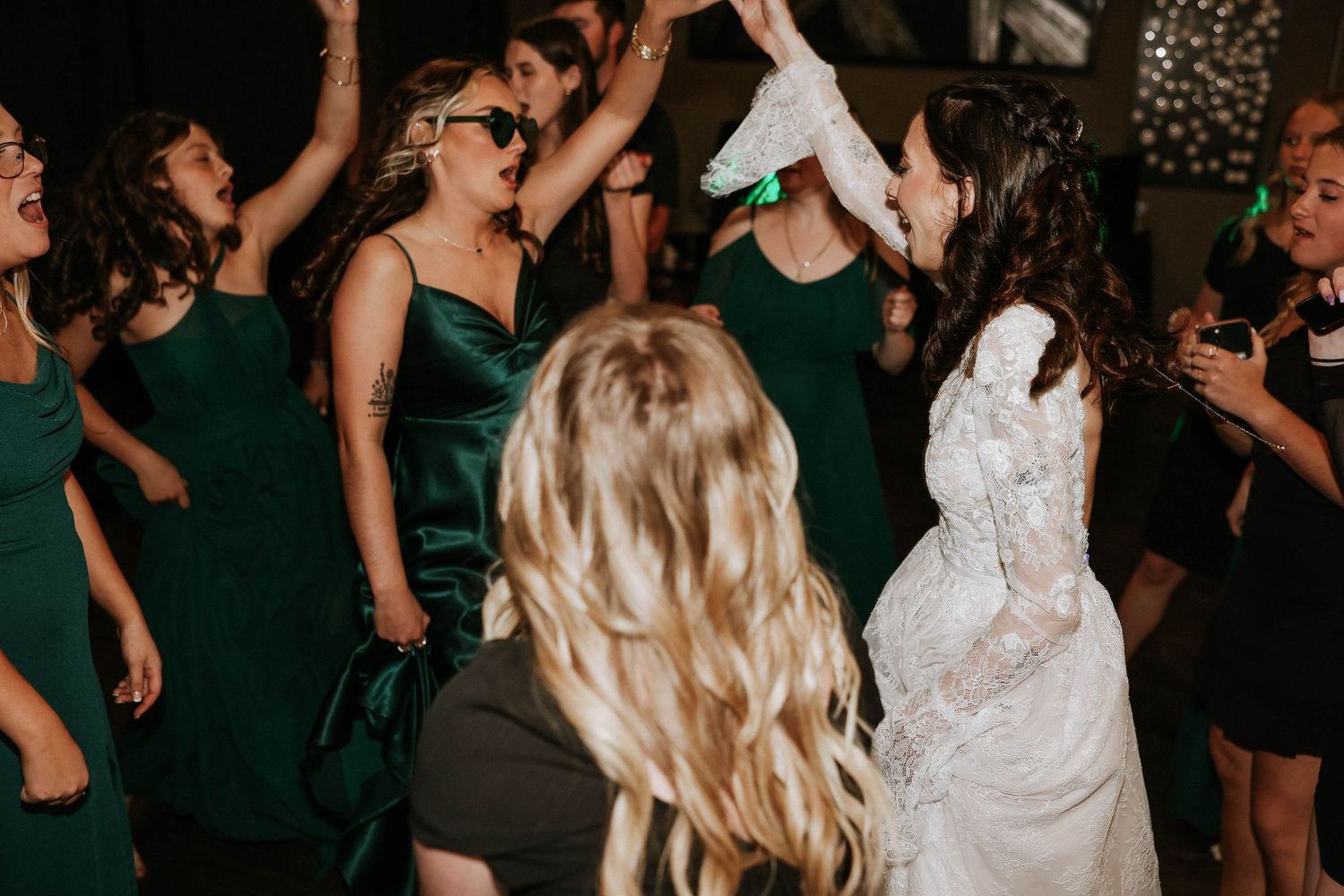 Bride in white lace gown dancing joyfully with bridesmaids in emerald green dresses at a lively wedding reception.