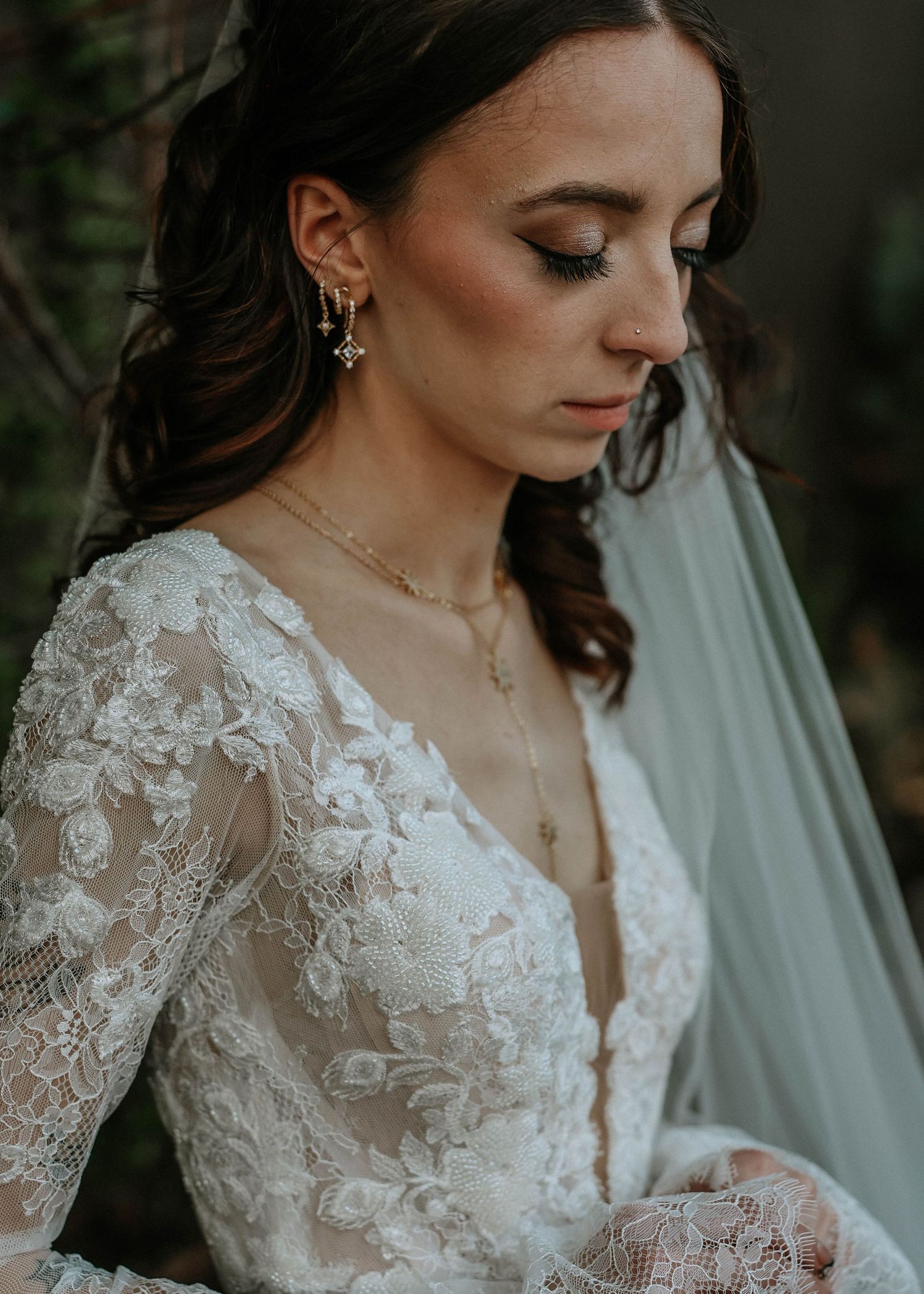 Bride in lace long-sleeve wedding gown with gold drop earrings and layered necklace, eyes closed, soft bridal makeup.