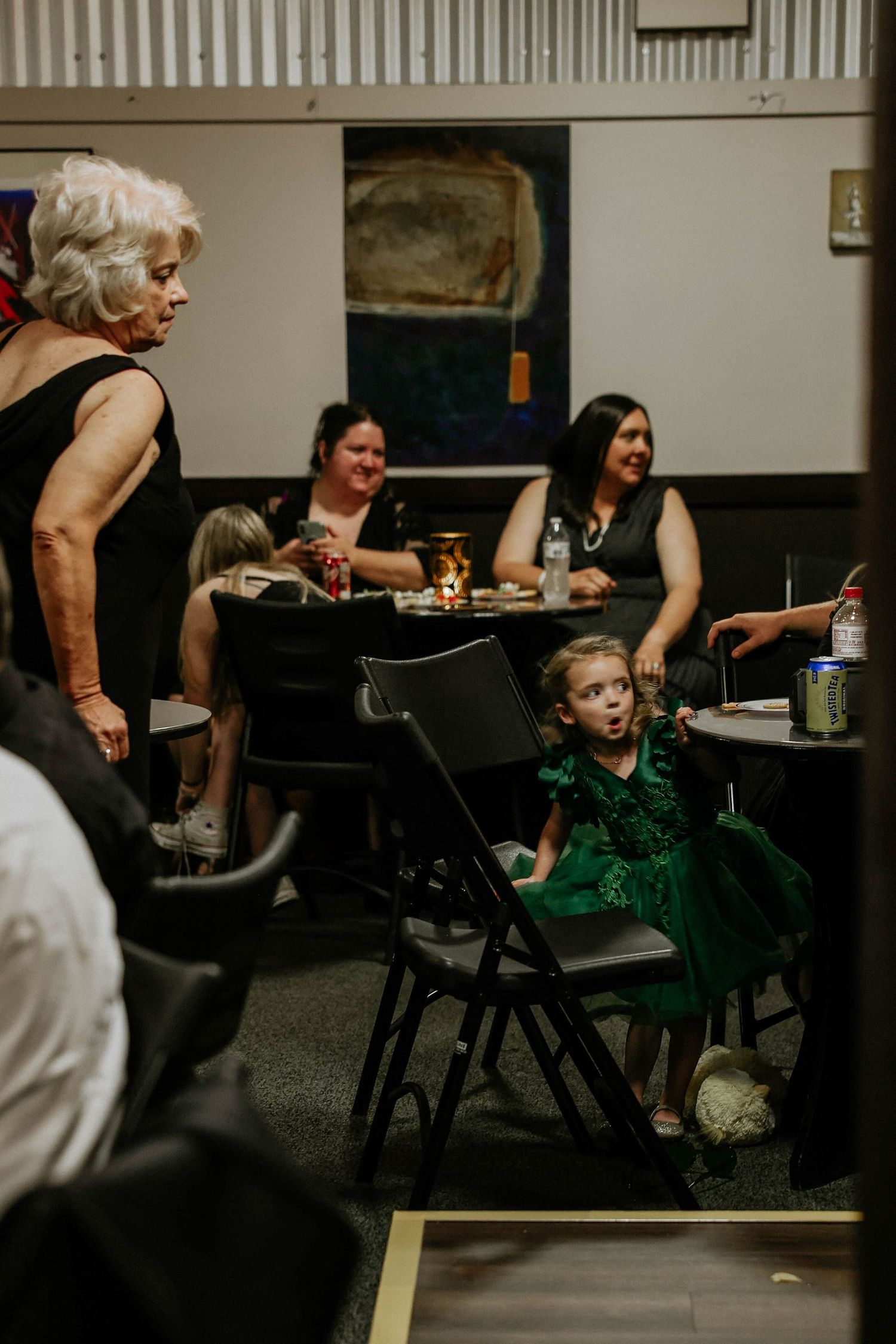 Young girl in green dress at indoor event with adults seated at tables in dimly lit venue.