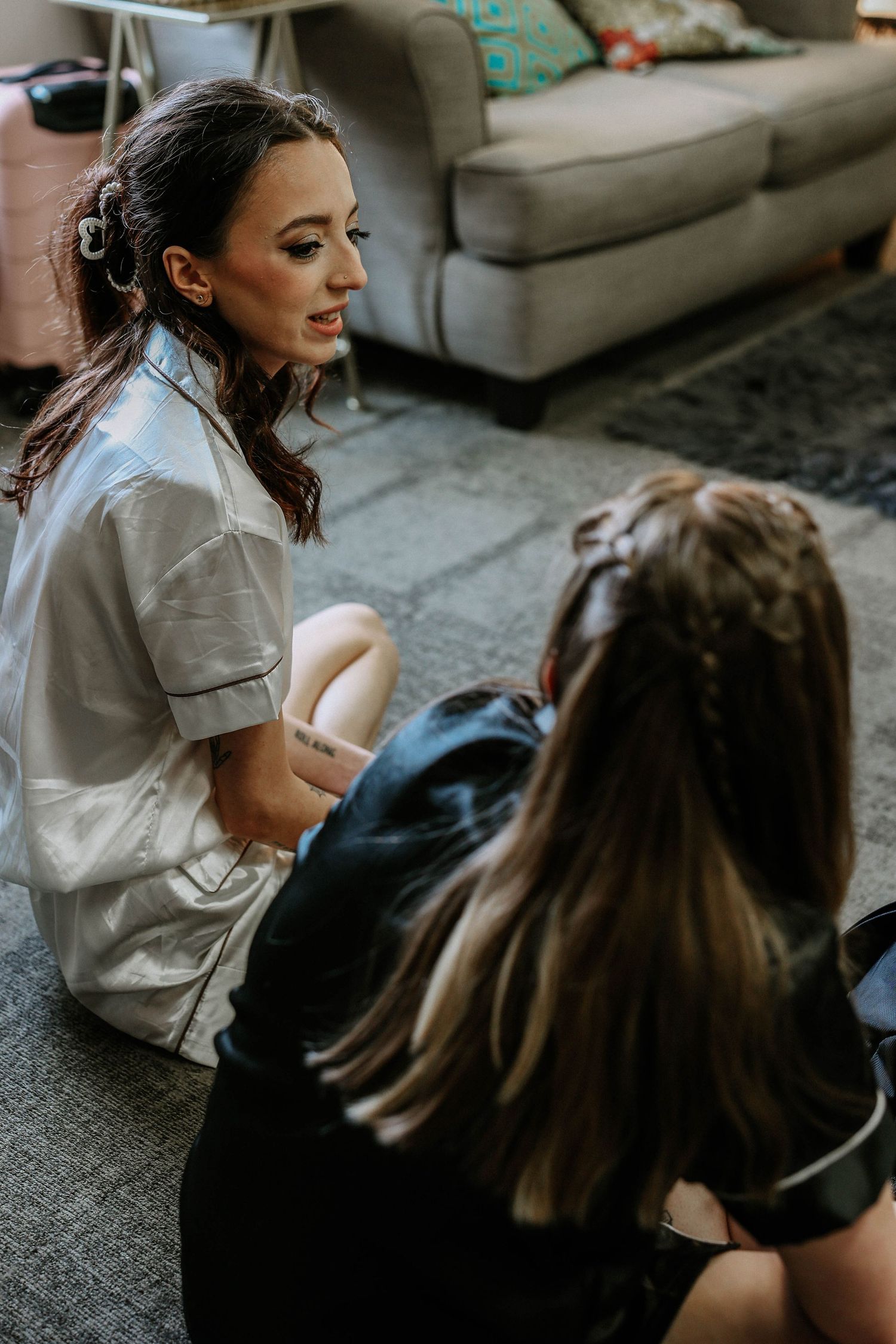 Bride in white satin robe sits on floor conversing with bridesmaid during wedding morning preparations in a hotel suite.