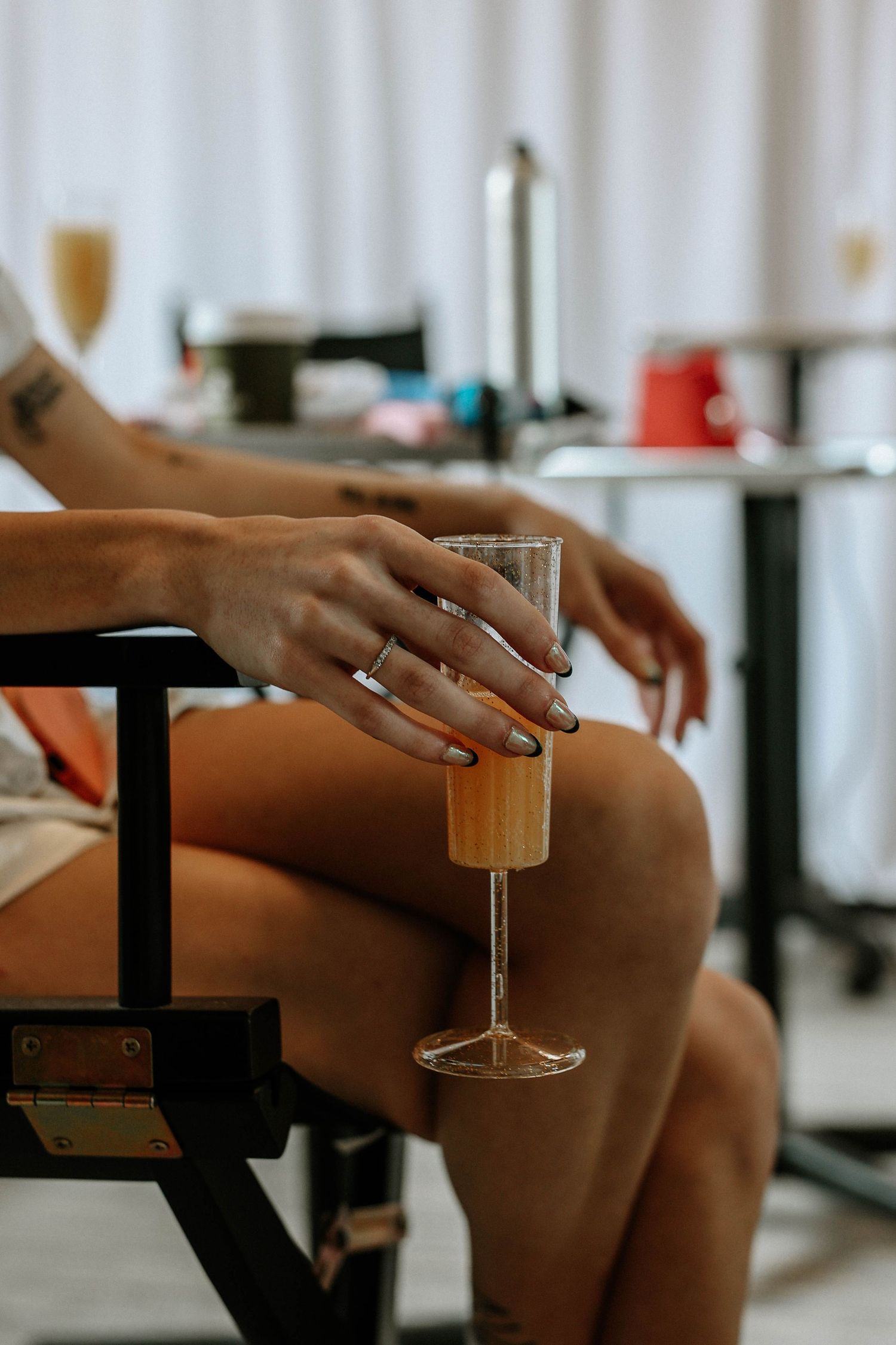 Woman with tattoo and rings holding a champagne flute on her lap while seated in a director's chair.
