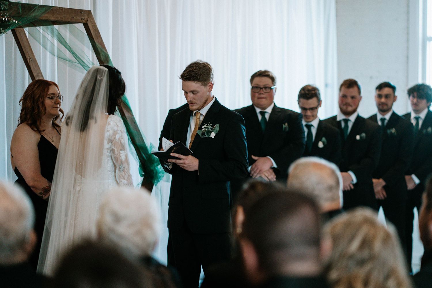 Groom reads vows at elegant indoor wedding ceremony with groomsmen standing behind him.