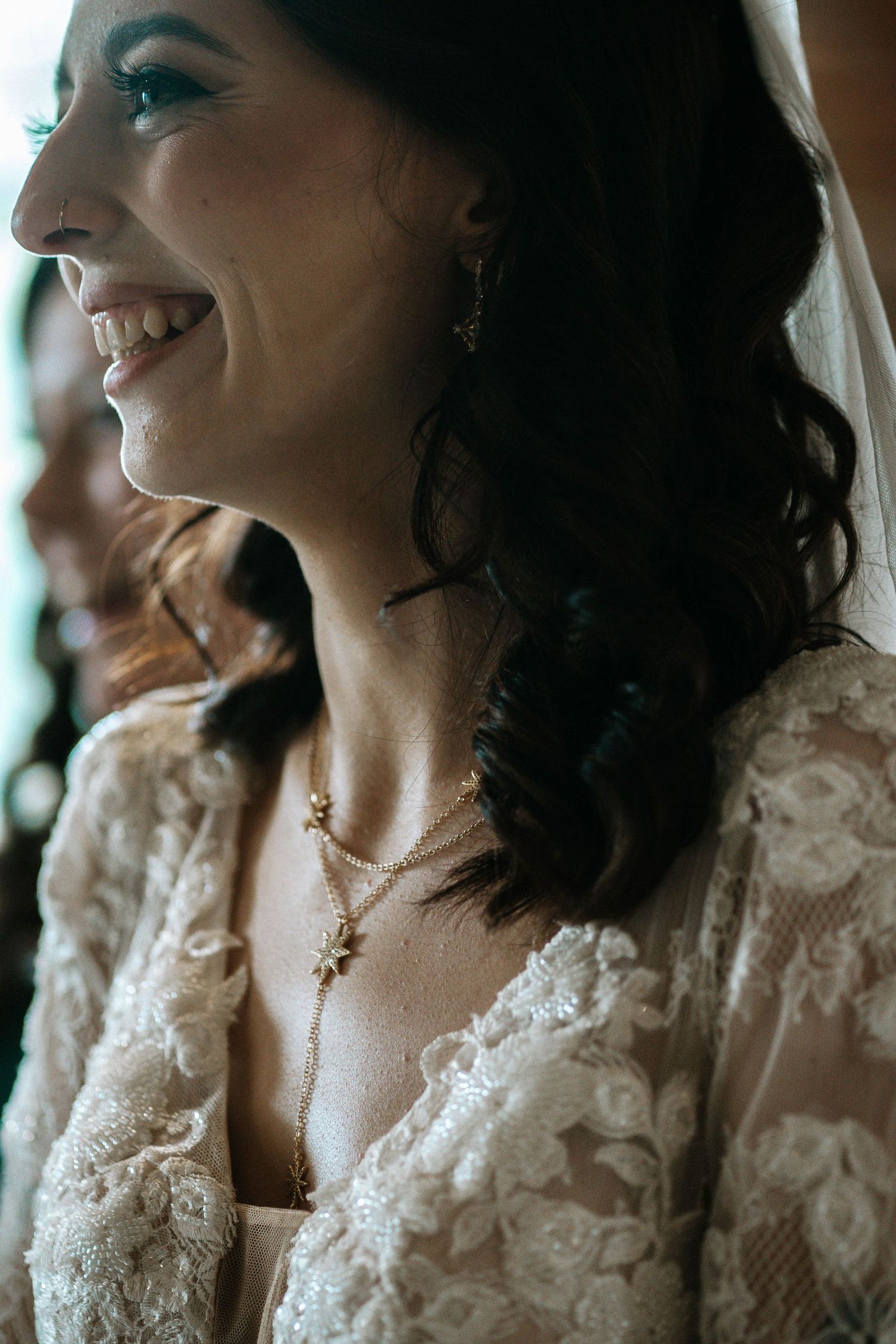 Smiling bride in lace wedding dress with gold necklace and earrings, wearing a veil, captured in a candid side profile.