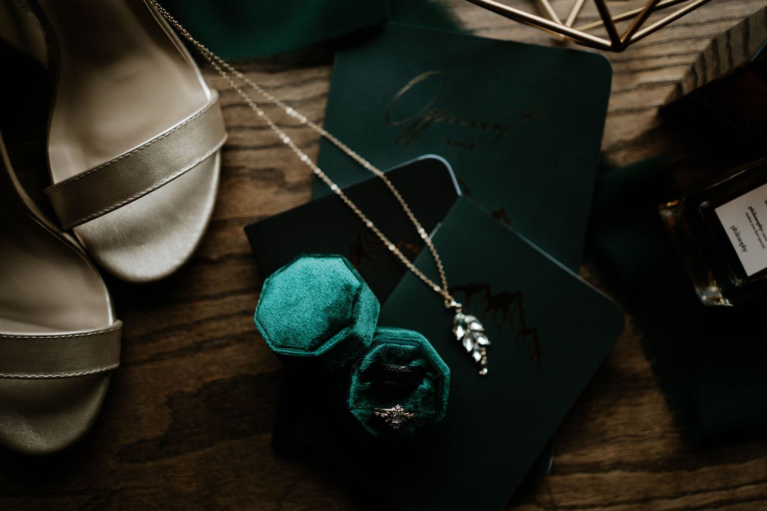 Elegant wedding flat lay with silver heels, green velvet ring box, diamond ring, delicate necklace, and stationery on wood.