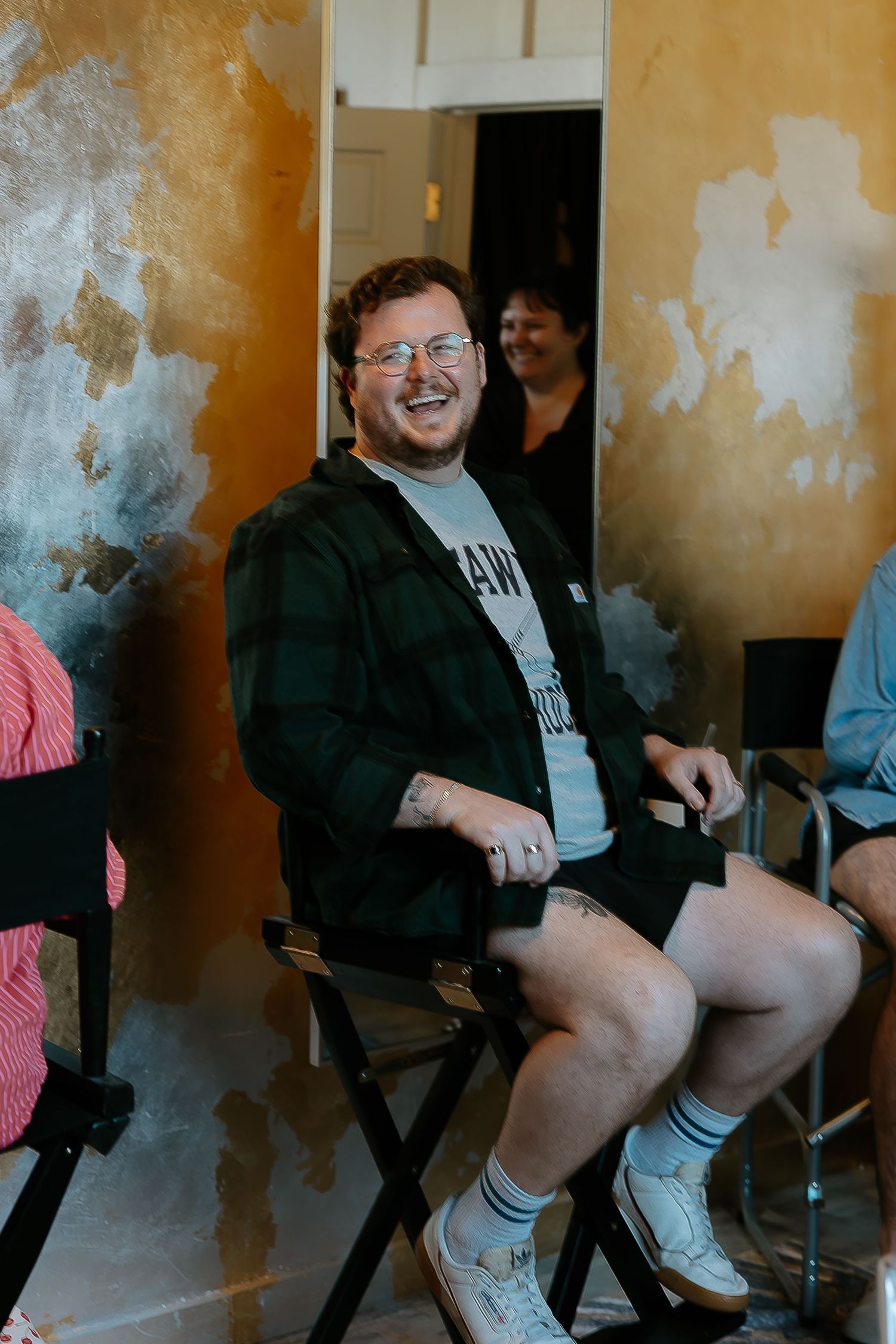 Smiling man in glasses and flannel shirt seated in a director's chair indoors.