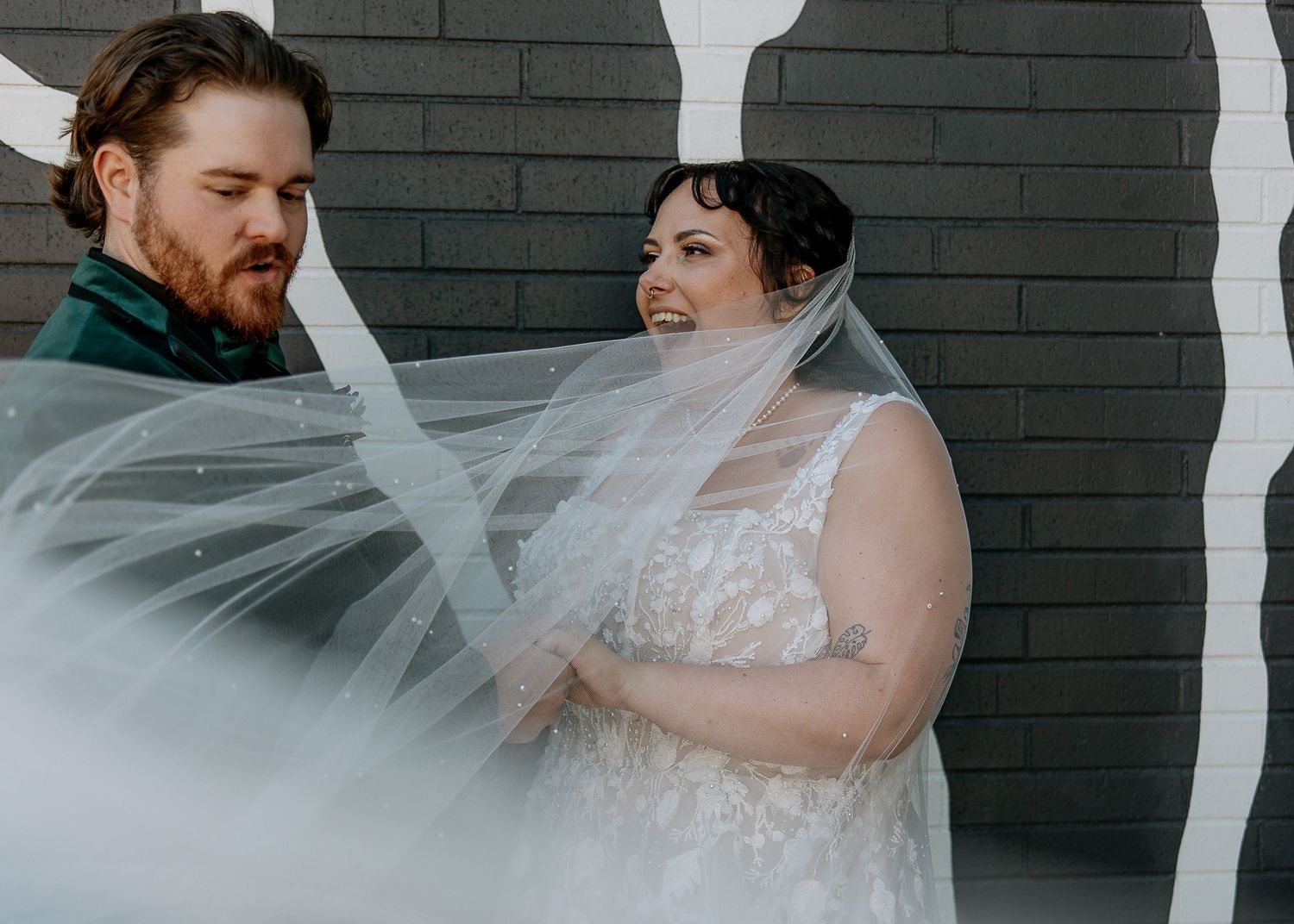 Bride in lace gown with flowing veil stands with bearded groom in green suit against dark brick wall.