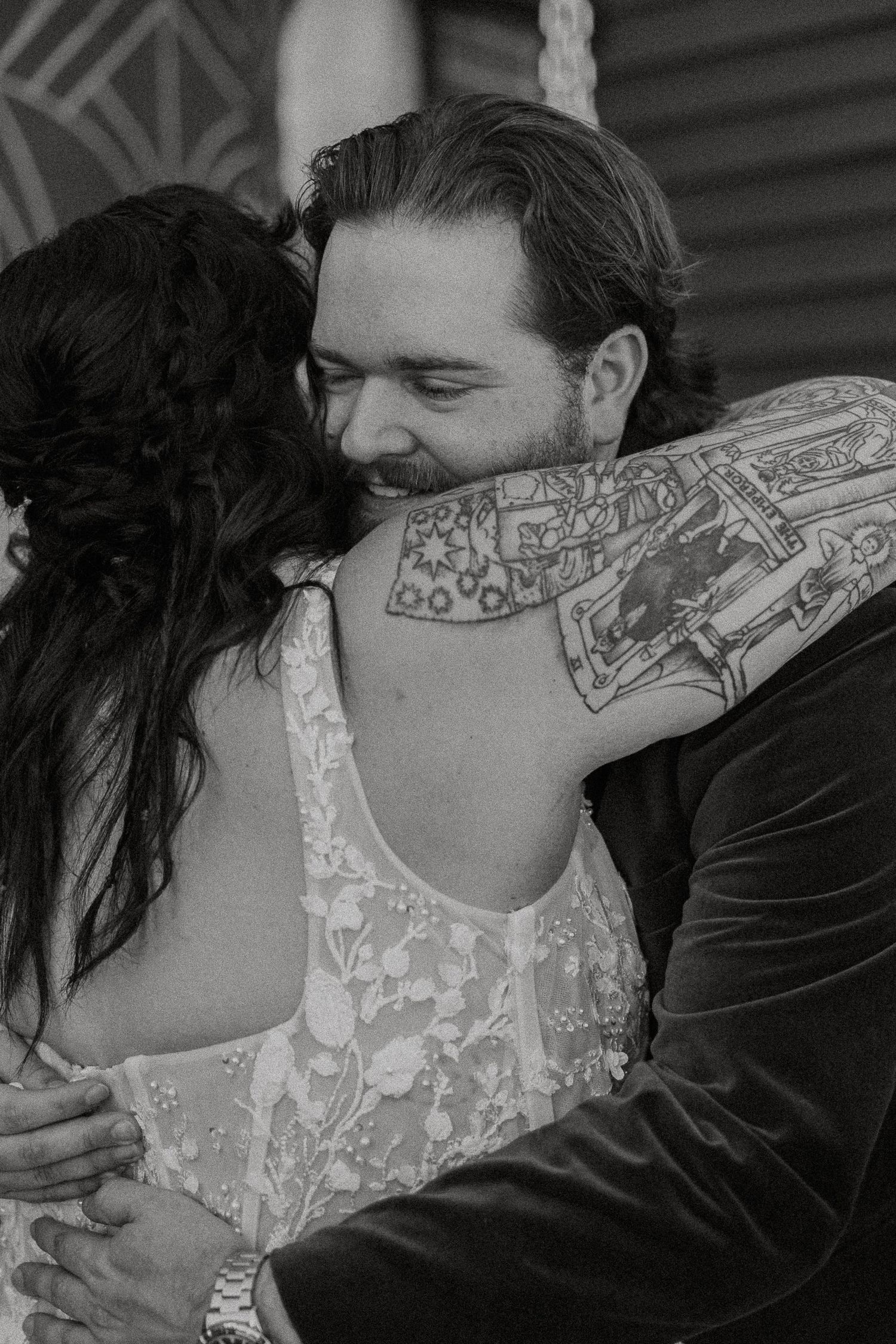 Black and white close-up of smiling groom embracing tattooed bride in lace wedding dress during first look.