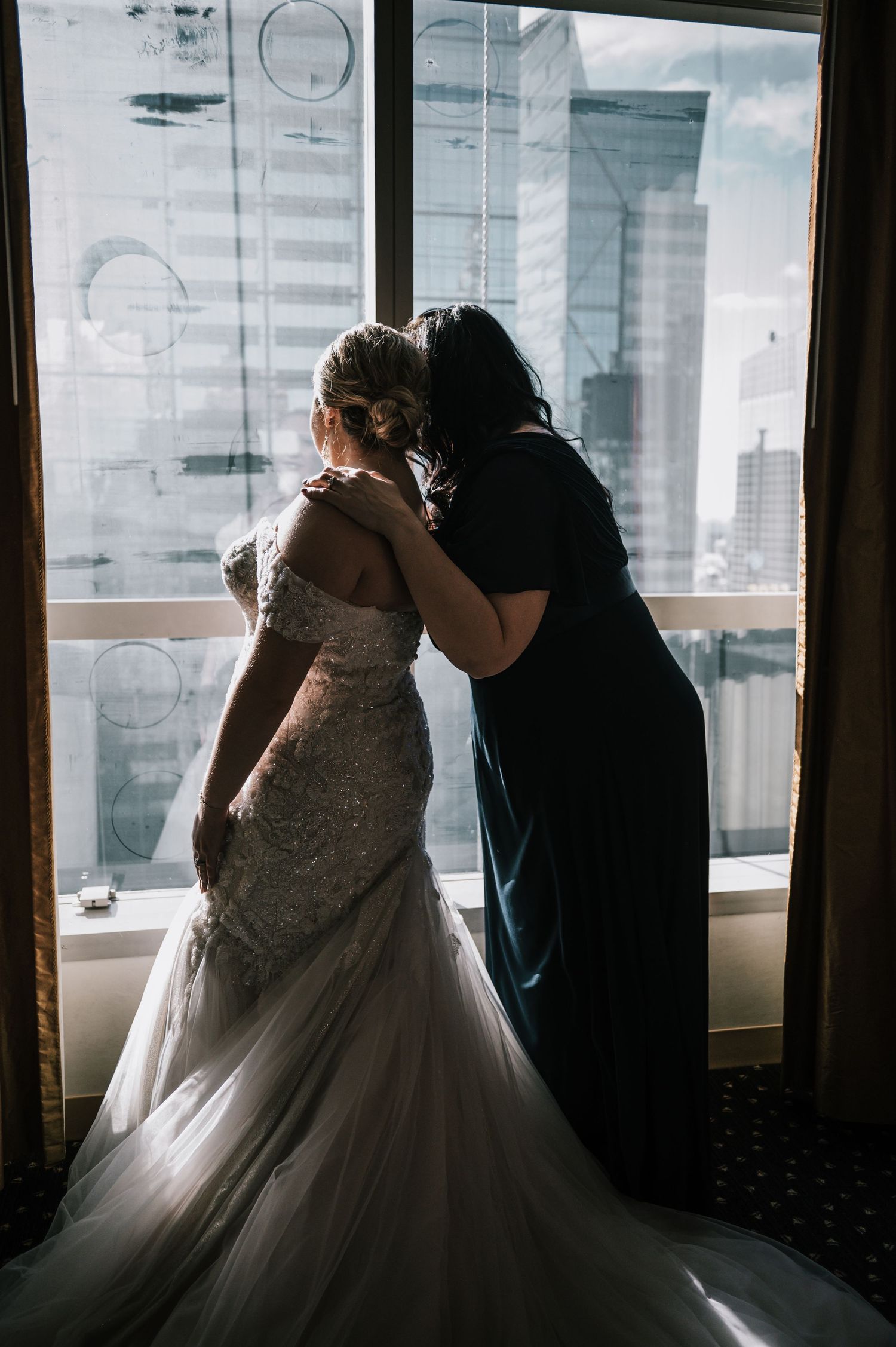 Bride in beaded mermaid gown shares tender moment with woman in blue dress by city window.