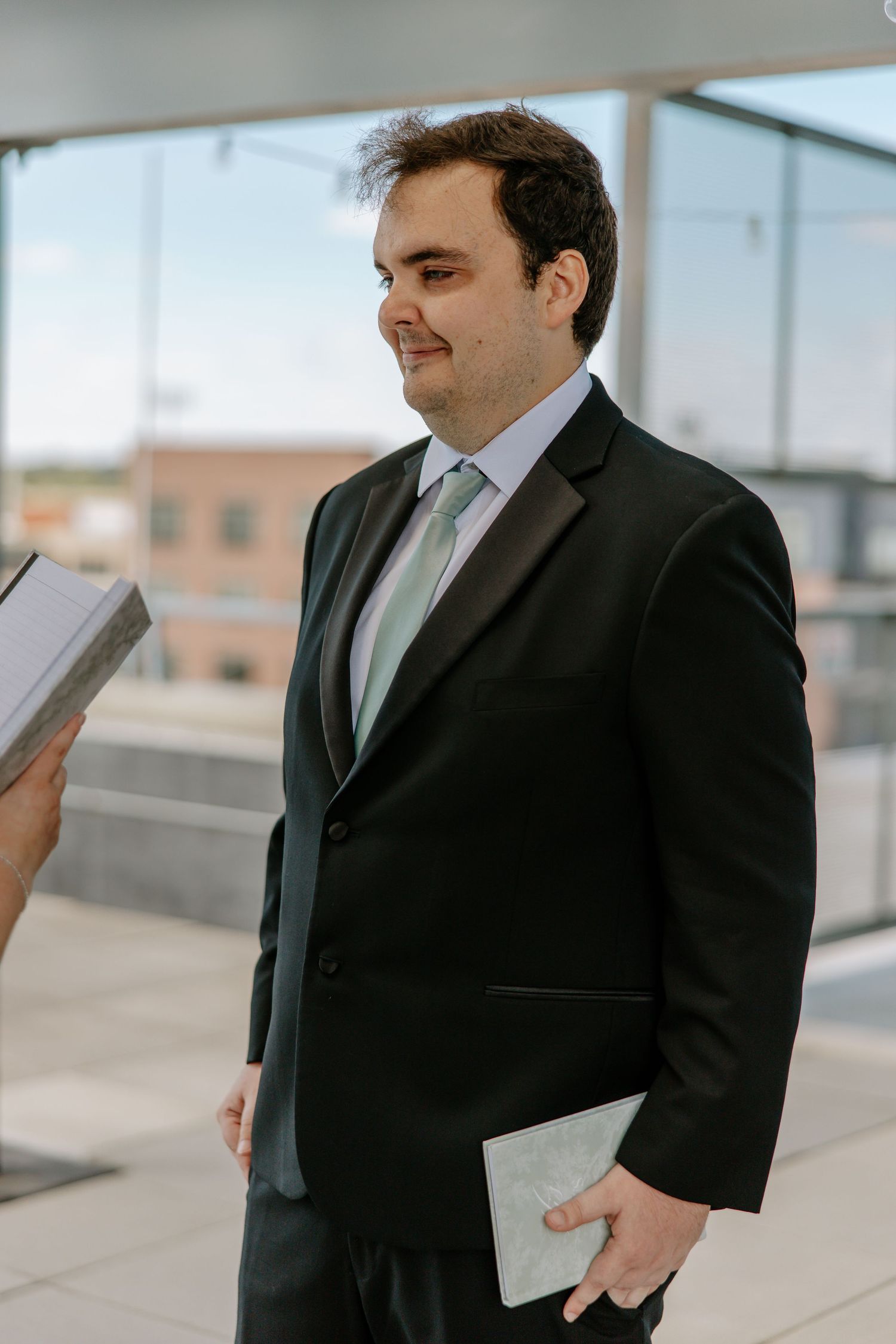 Groom in black tuxedo holds vow book while standing on rooftop during intimate wedding ceremony.