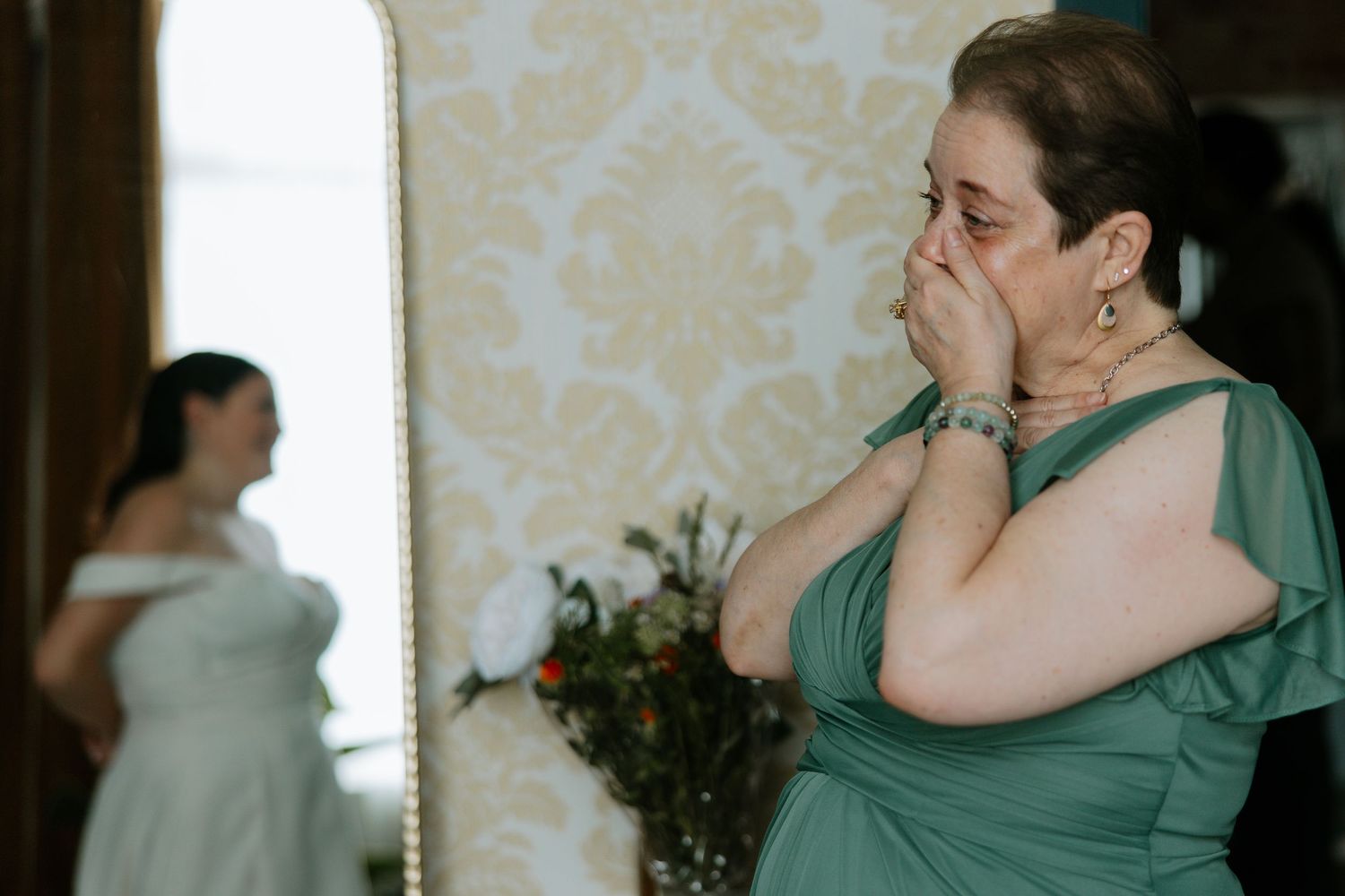 Emotional mother of the bride in green dress crying while bride is reflected in mirror.