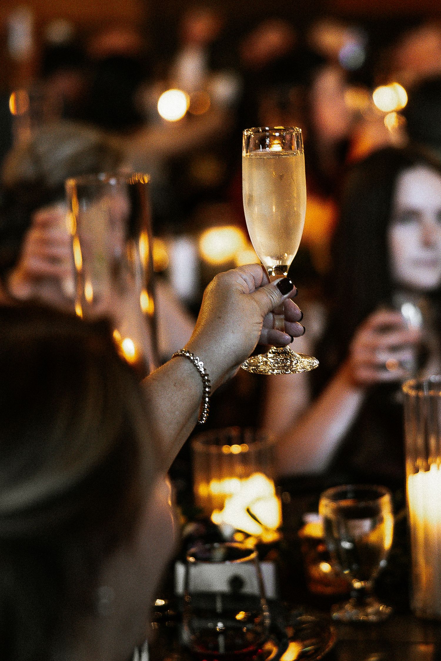 A hand raises a champagne flute in a toast at a warmly lit wedding reception celebration.